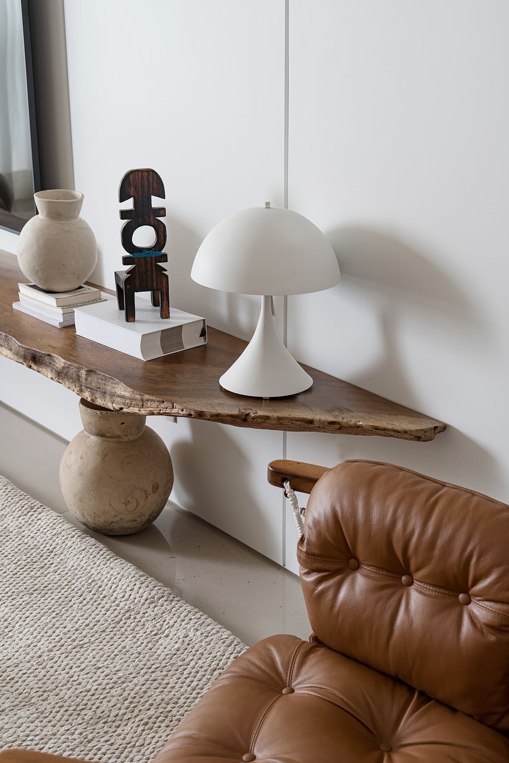 Detail: clay vessels, a carved sculpture and a white mushroom lamp on a raw timber shelf above a tufted leather chair.