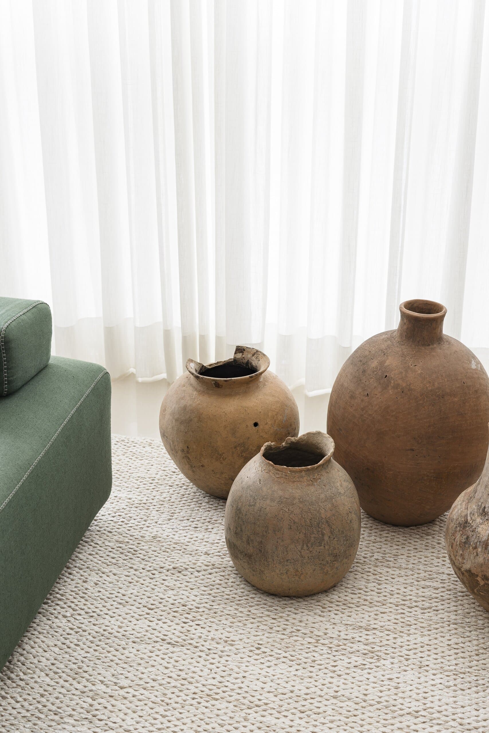 Detail: three large clay pots on a woven rug beside the green sofa, sheer curtains behind.