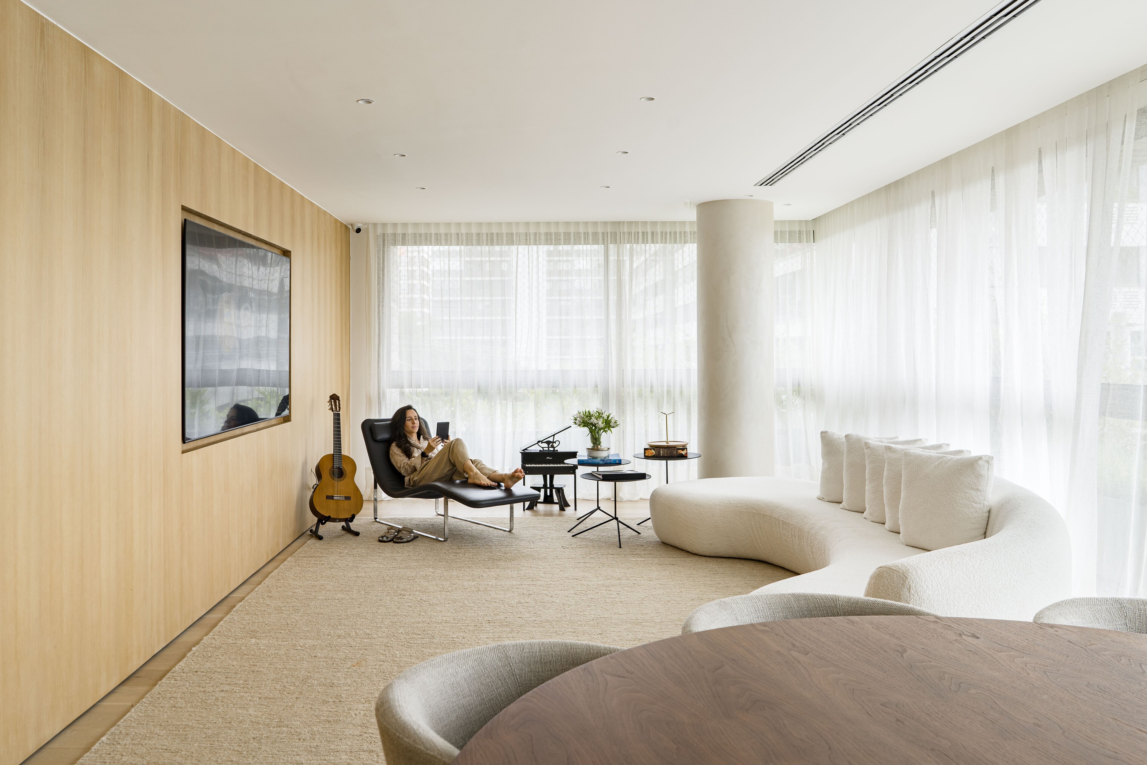 A pale living room with oak-panelled walls and sheer curtains, a woman in a black lounger beside a guitar and a curved white sofa around a round column.
