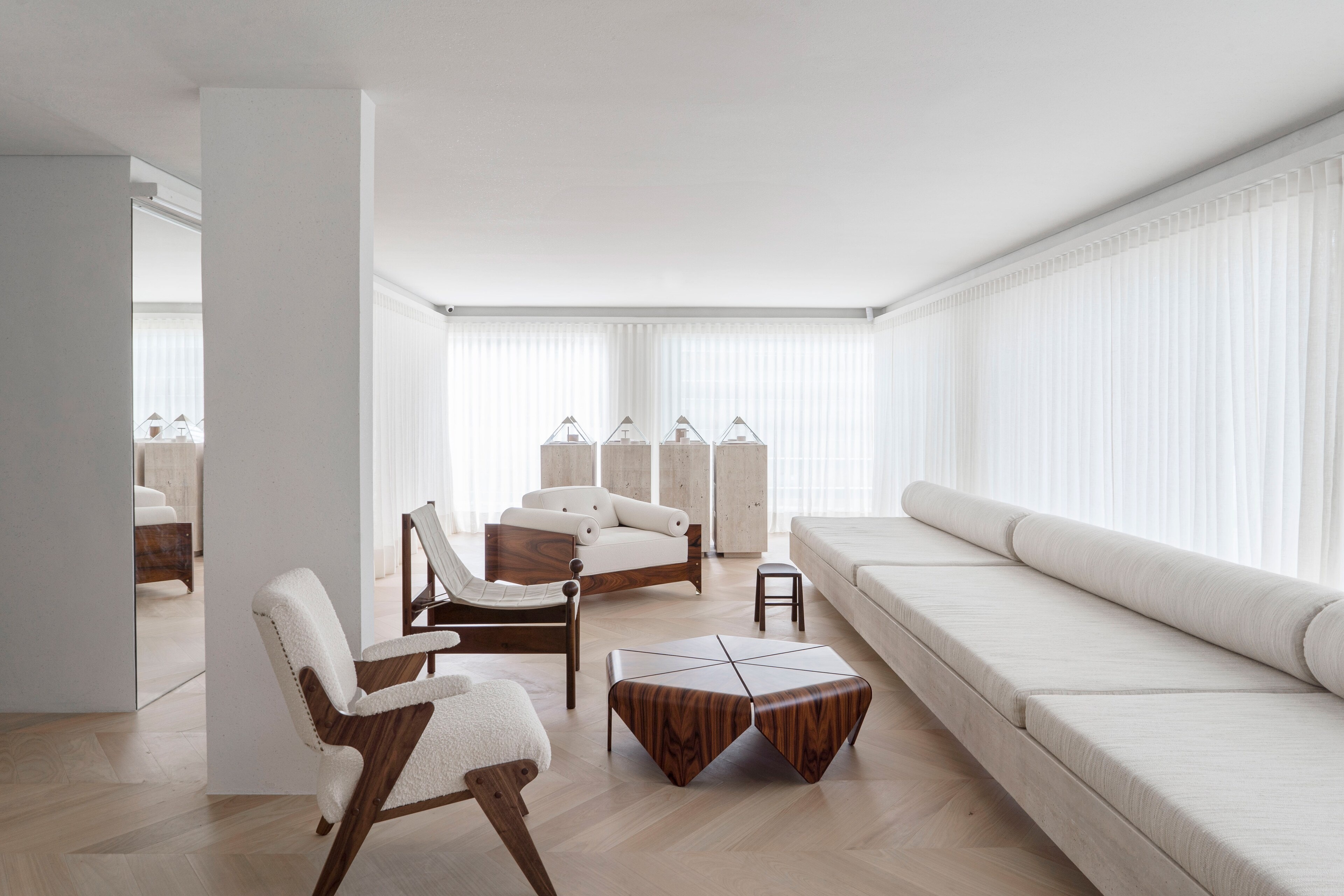 A pale living room behind sheer curtains: a long white bench sofa, a faceted walnut coffee table, bouclé and timber armchairs and travertine plinths beyond a white column.