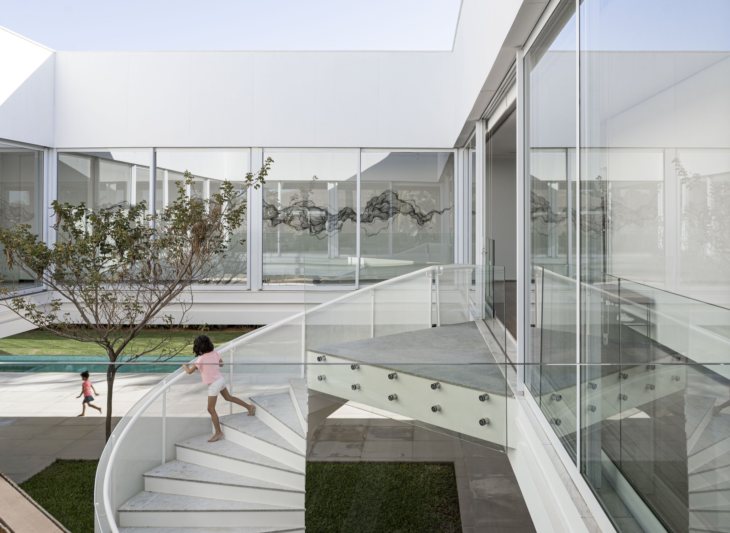 A white glass courtyard with a spiral stair and a slender tree, a child running past the pool's edge.