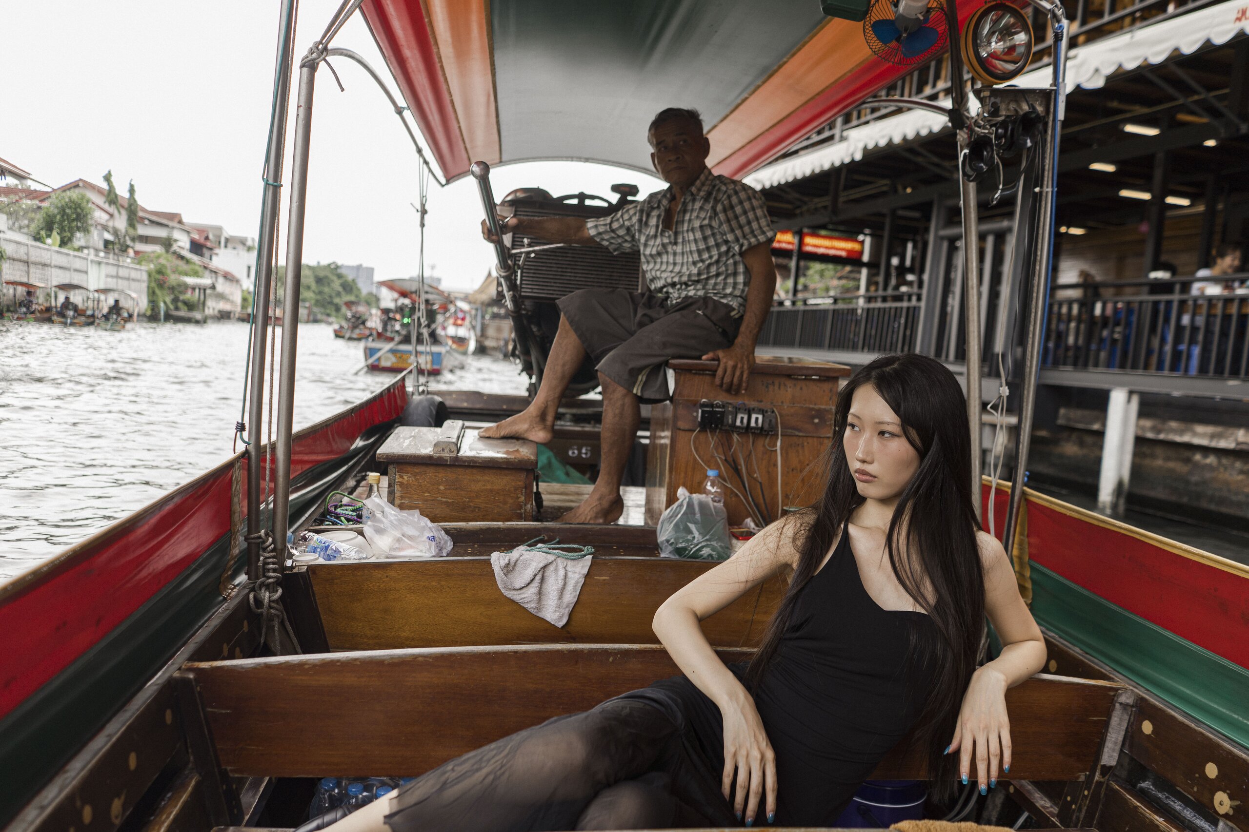 A woman in a black dress reclines on a wooden bench of a painted longtail boat, the boatman steering behind her on the river.