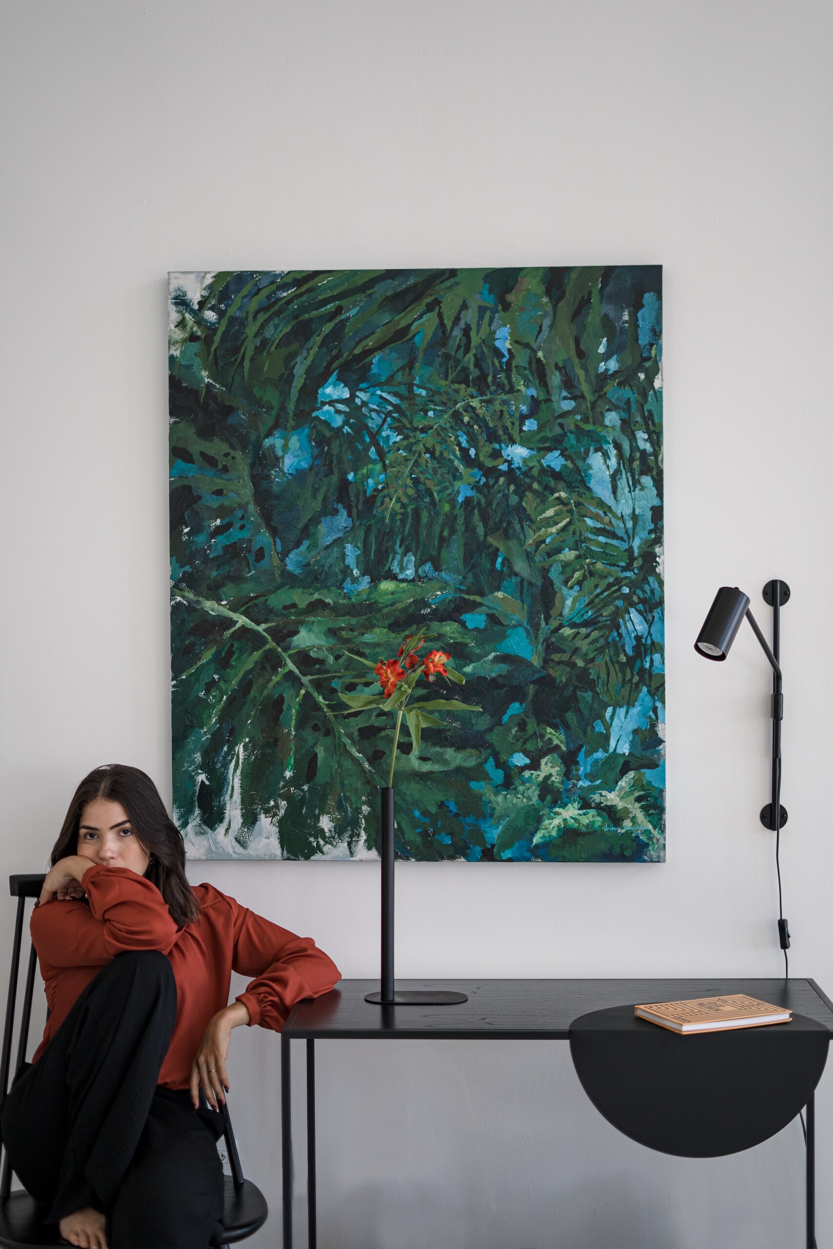 A woman in a rust jumper sits beside a black metal desk with a half-moon shelf, a jungle painting on the white wall above.