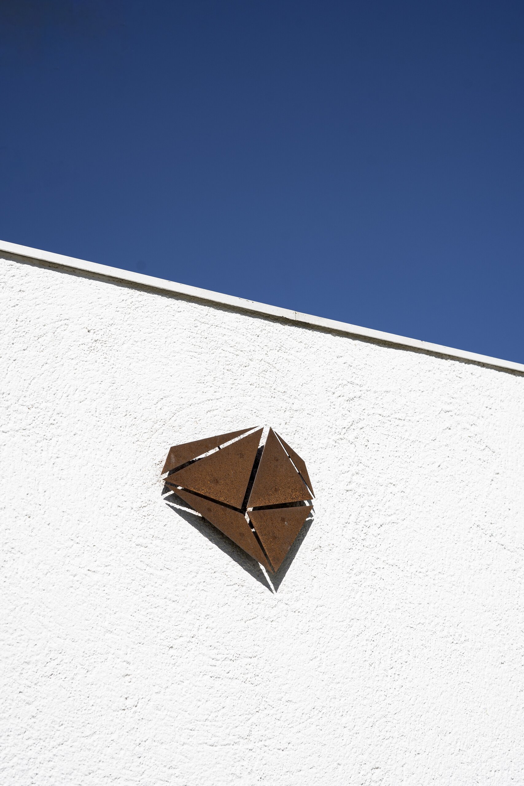 A faceted rusted-metal gem fixed to a white rendered wall under a deep blue sky.