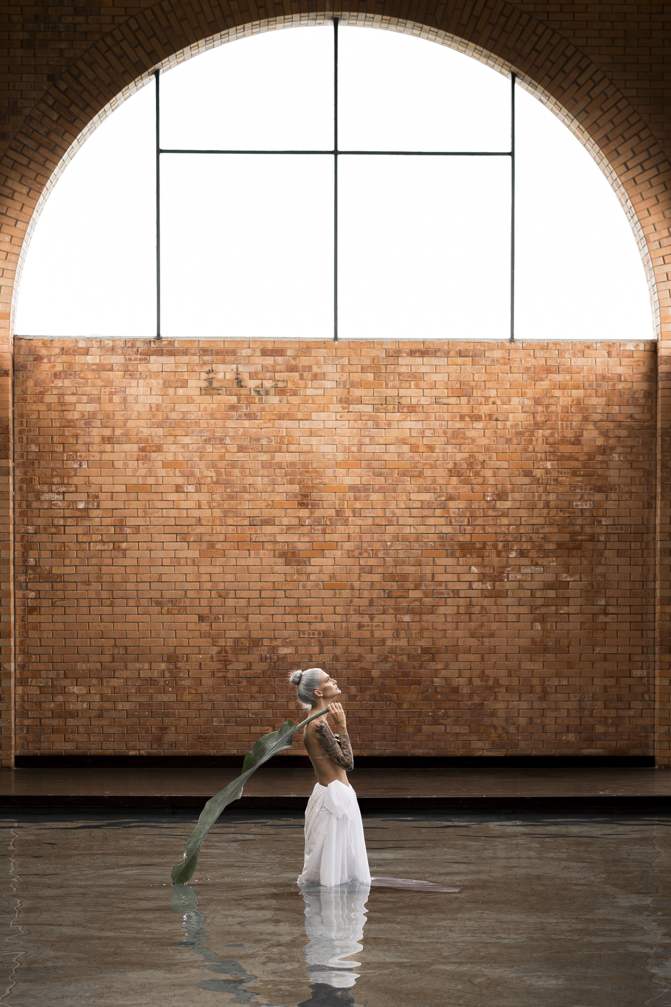 Beneath a tall arched window, a woman in a white skirt stands in shallow water against a brick wall, a long leaf over her shoulder.
