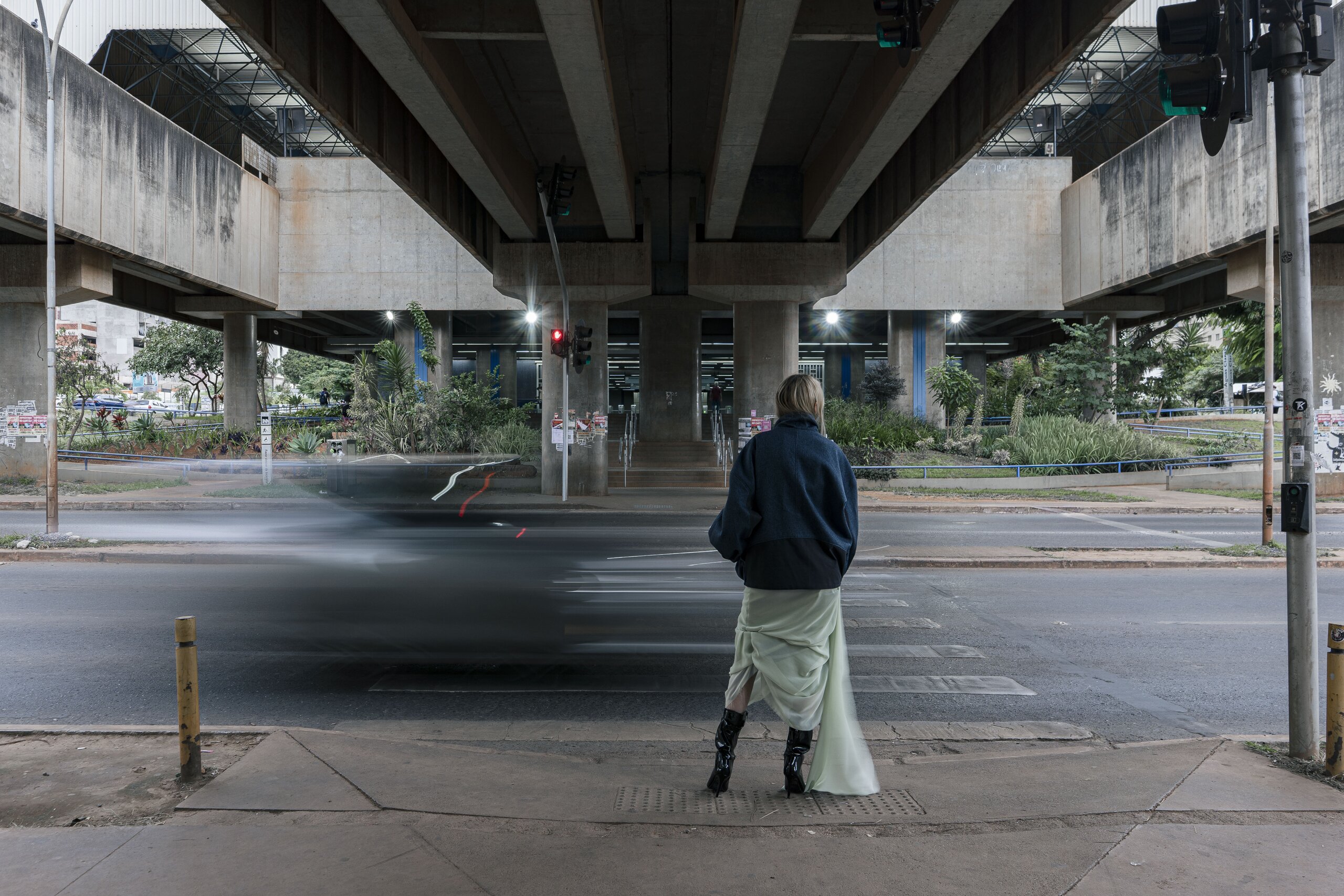 Seen from behind, a woman in a navy jacket and pale skirt waits at a crossing beneath a concrete overpass as a car streaks past.