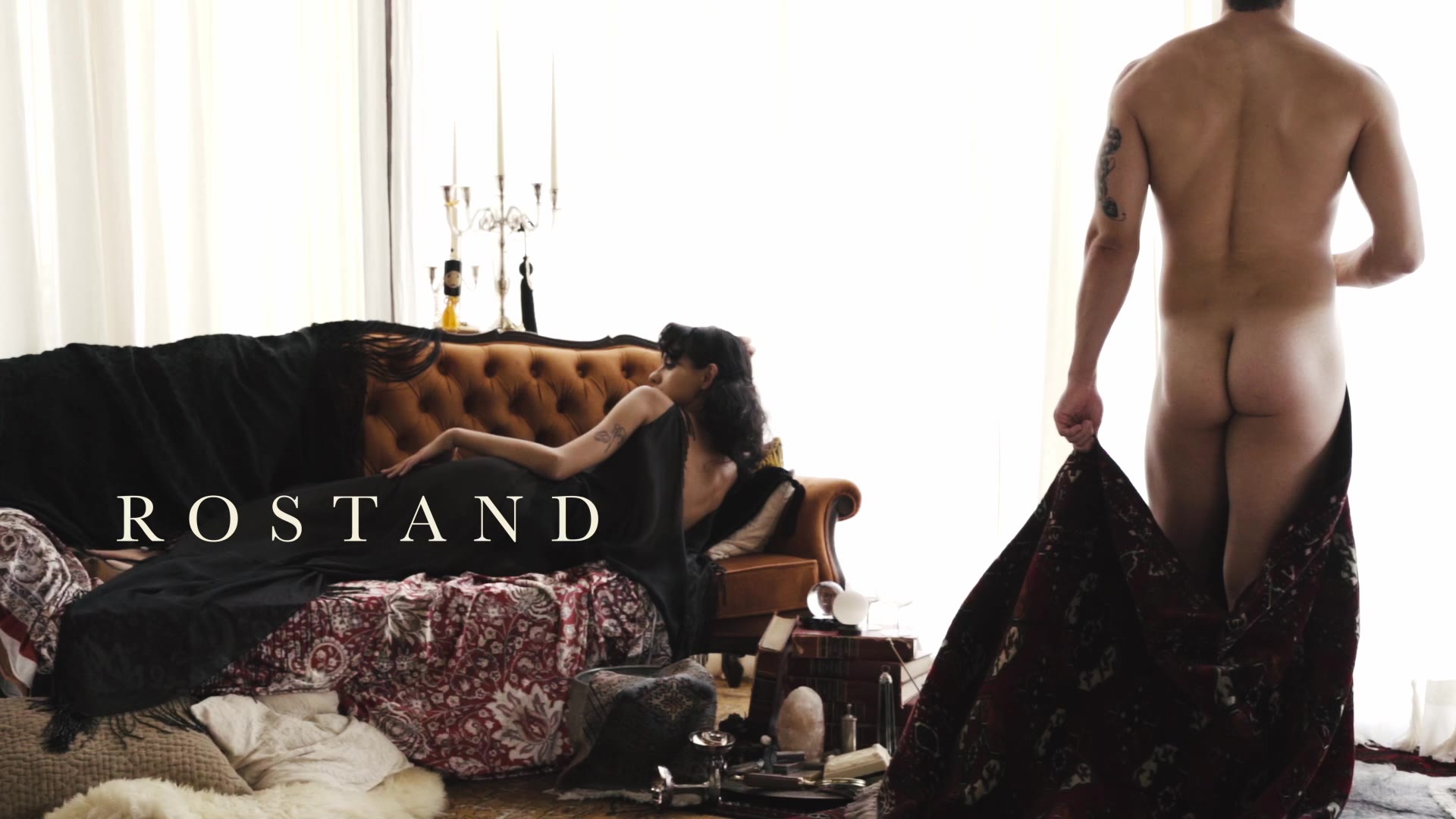In a bright room with a candelabra and a leather sofa, a man seen from behind holds up a dark patterned cloth, the word ROSTAND across the frame.