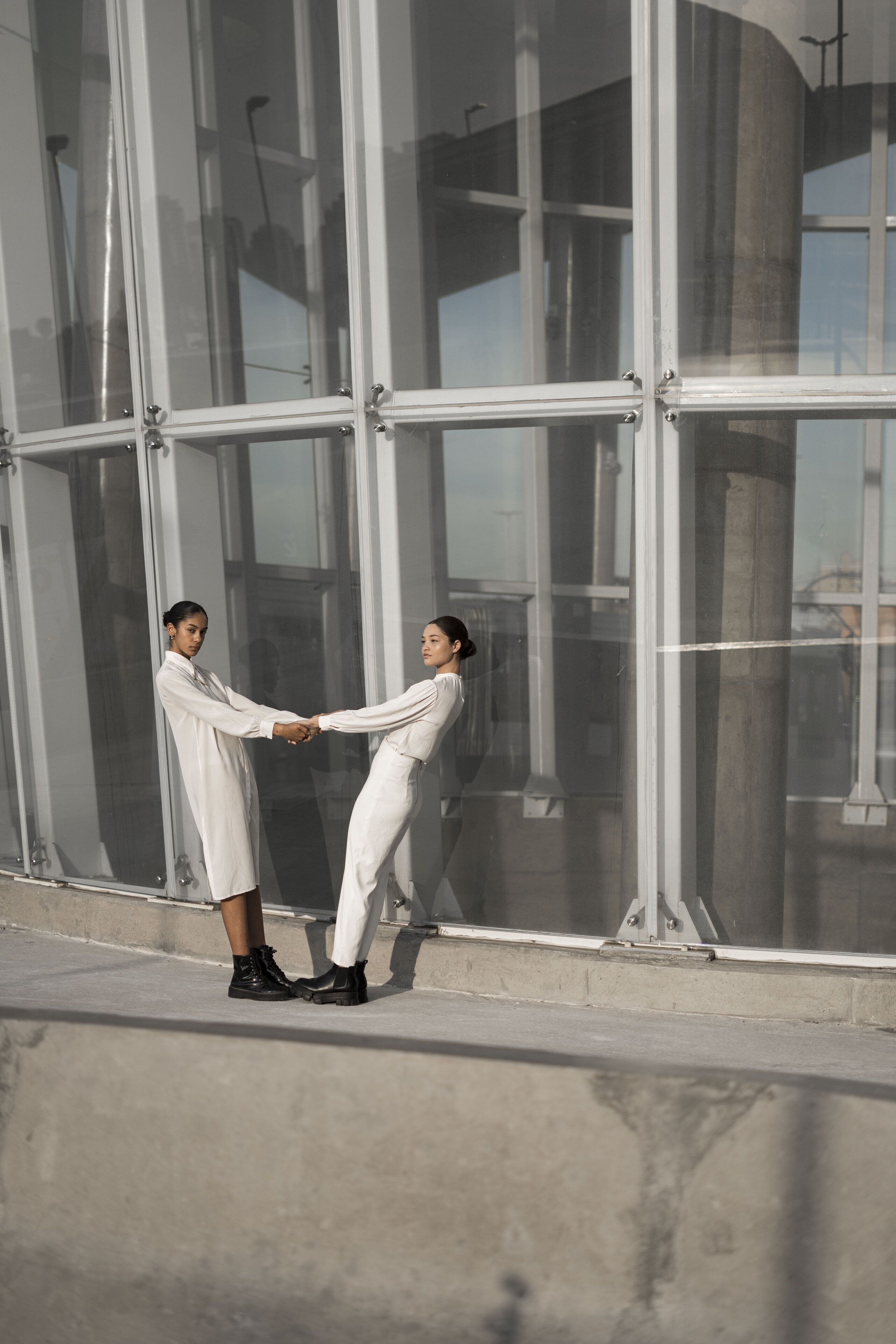 Two women in white hold hands at arm's length in front of a tall steel-framed glass facade.