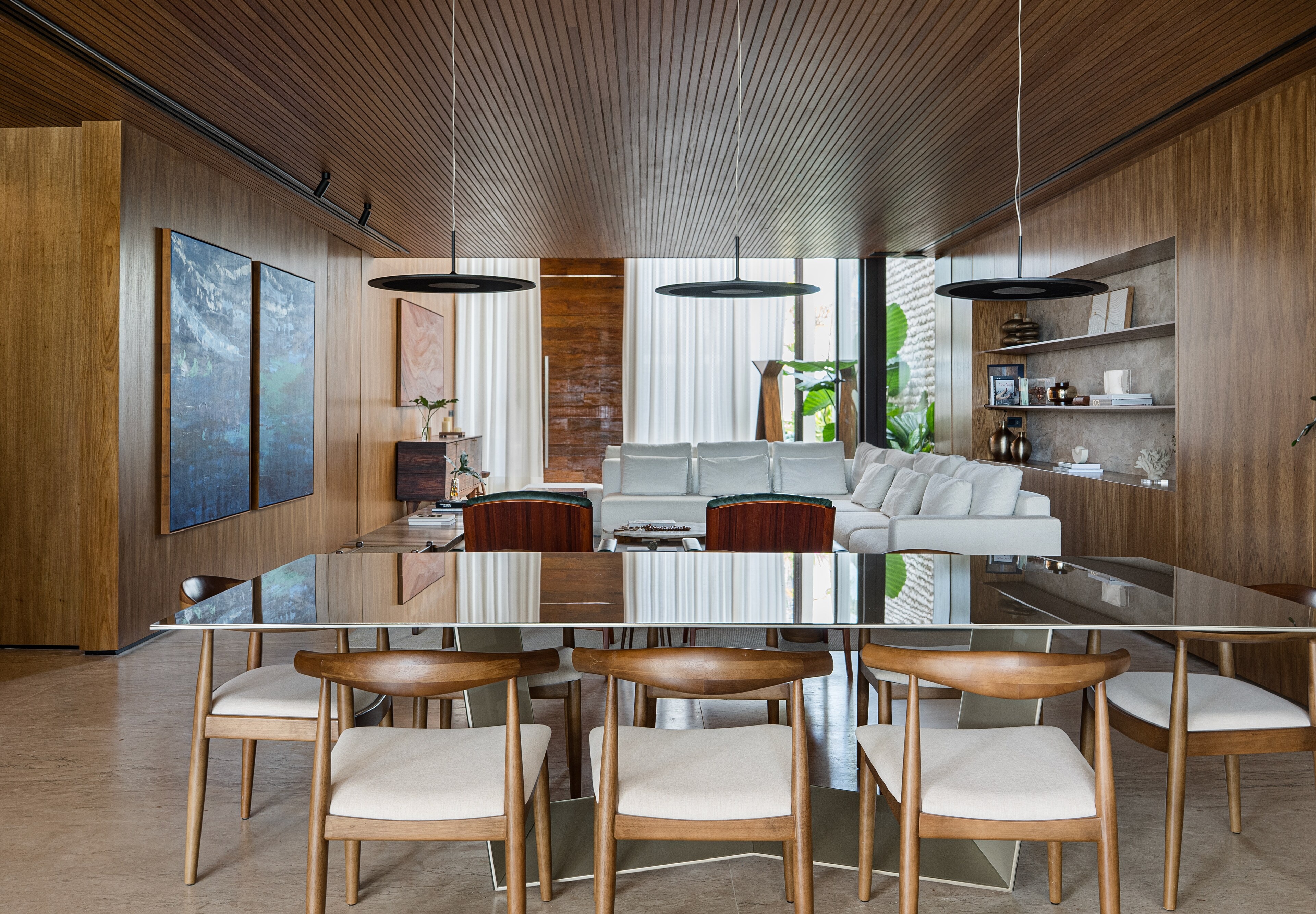 A long dining table with timber chairs under a slatted timber ceiling and black pendant lamps, the living room and blue paintings beyond.