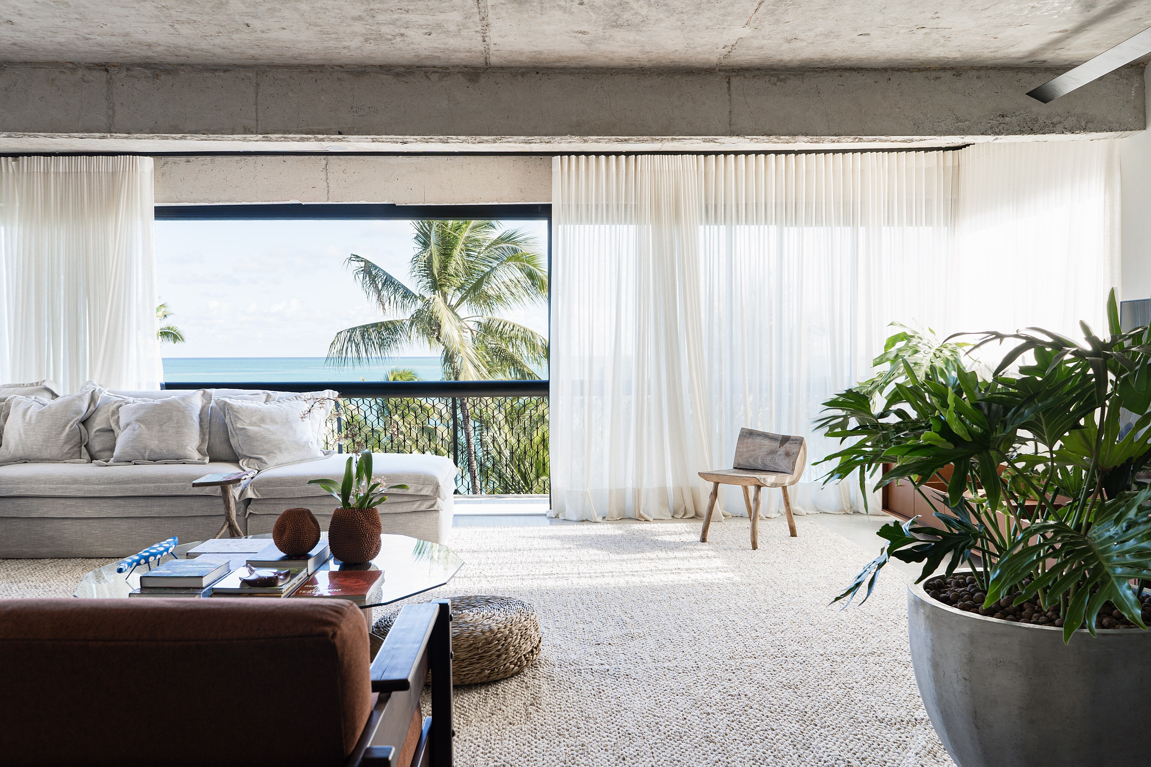 Uma sala sob a laje de concreto aparente: sofá claro, cortinas translúcidas e uma janela para os coqueiros e o mar.