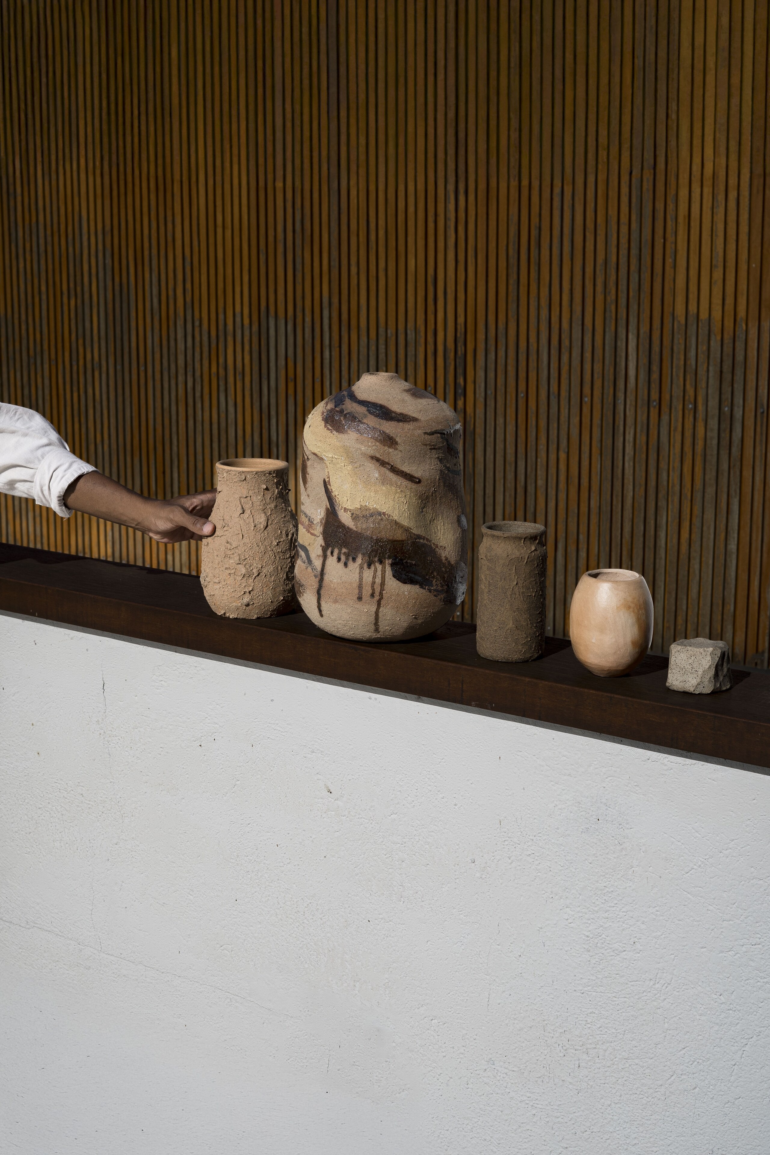 Hand-built ceramic vessels in a row on a dark timber ledge against slatted wood, a hand reaching in from the left.