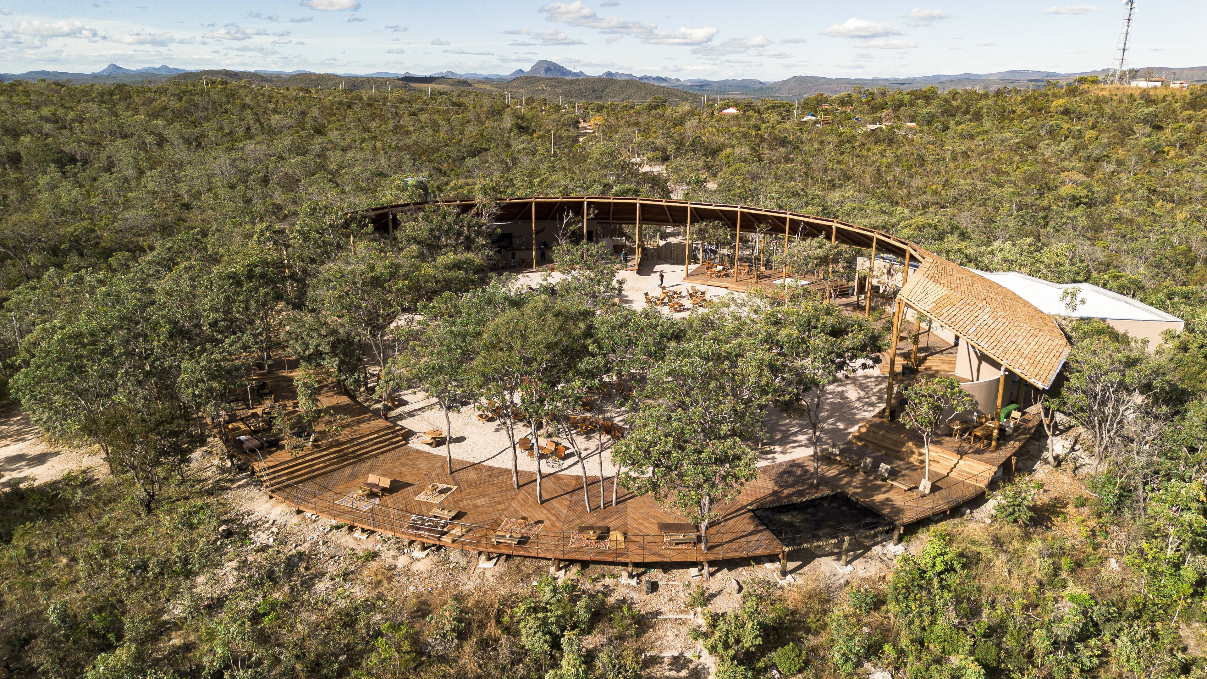 From the air: a ring of thatched roof around an open courtyard of trees, set on a rocky hilltop in the scrubland.