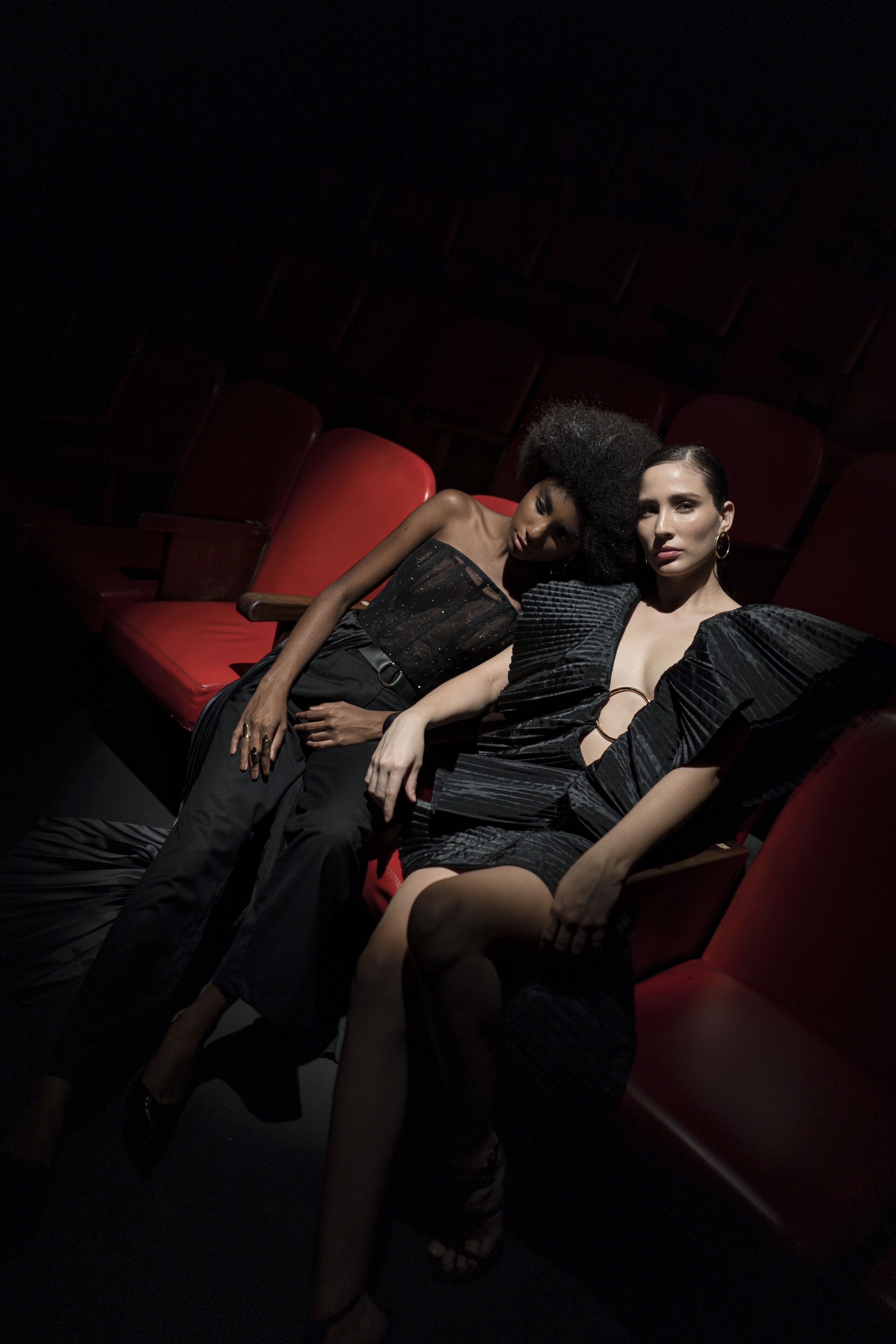 Two women in black reclining across red theatre seats, lit from one side in the dark.