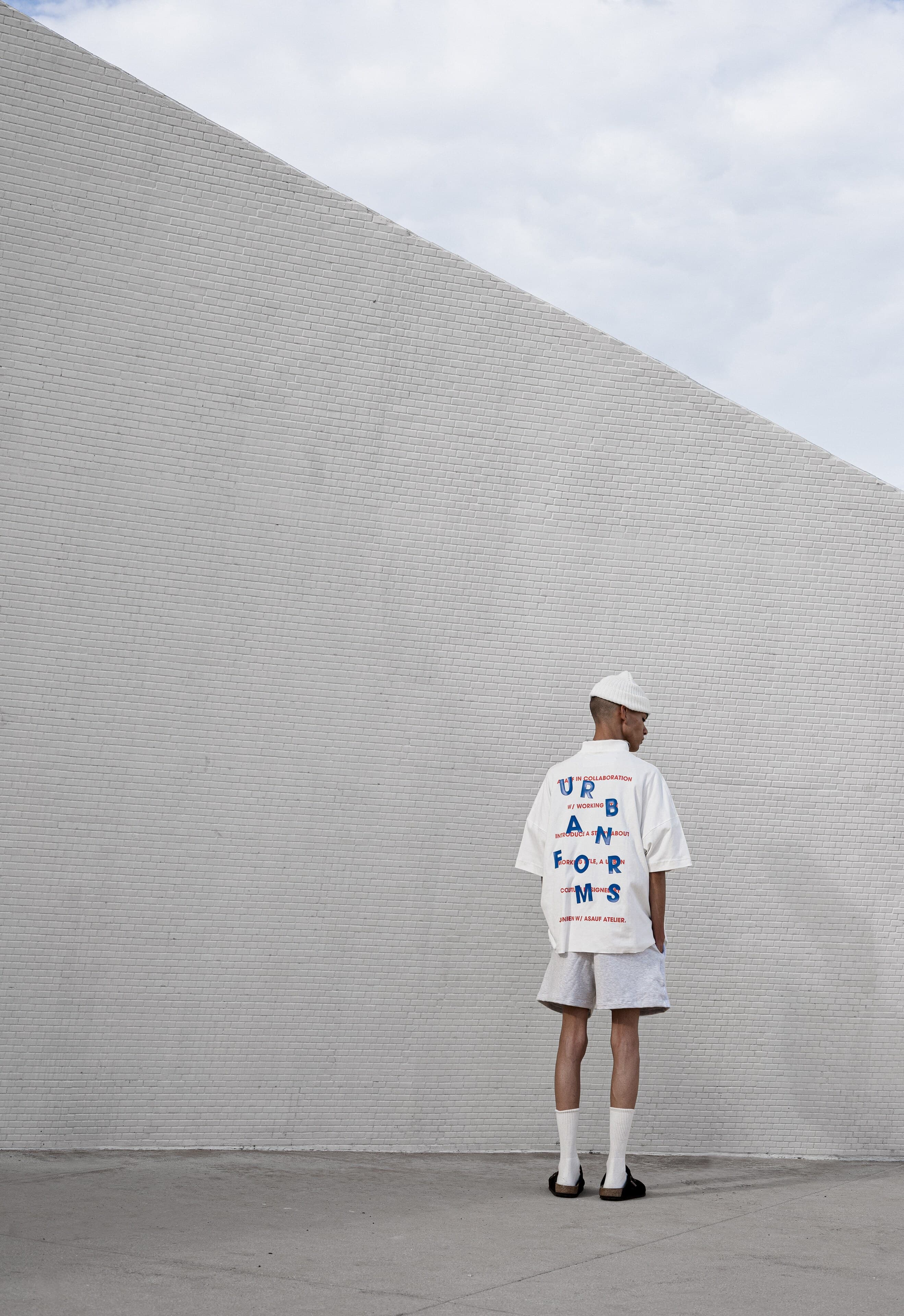 A man in a white cap and white shirt printed with blue lettering stands with his back to a tall white tiled wall under a cloudy sky.
