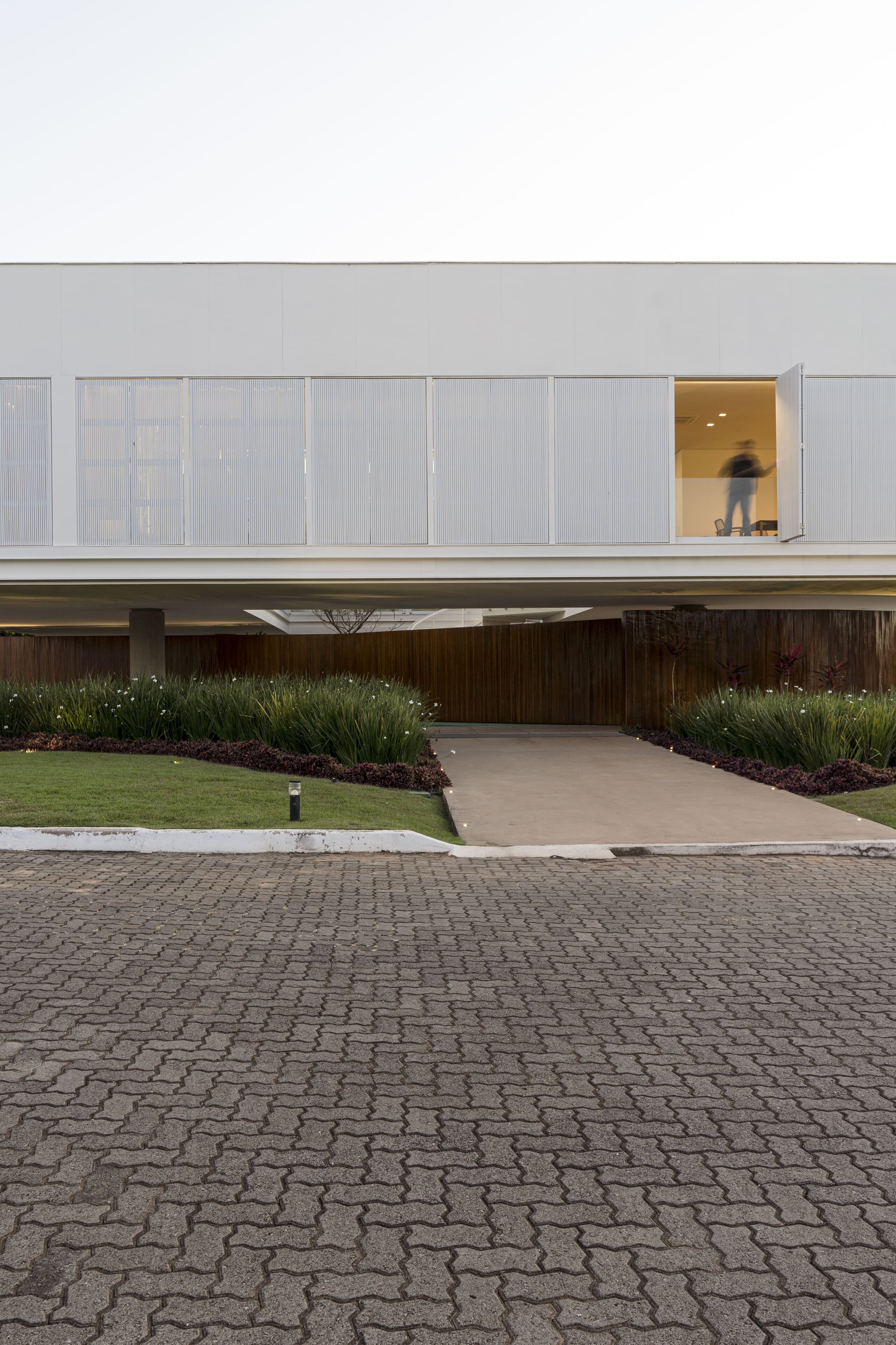 A long white box floating above a timber base, a lone figure in its one lit window, brick paving in the foreground.
