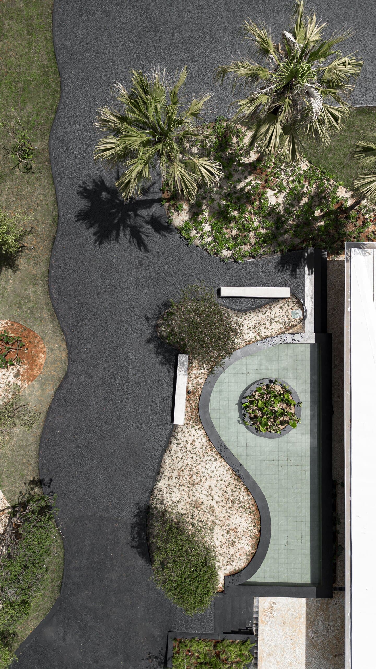 From above: a dark cobbled path winding past palms and a curved concrete bench around a round planter.