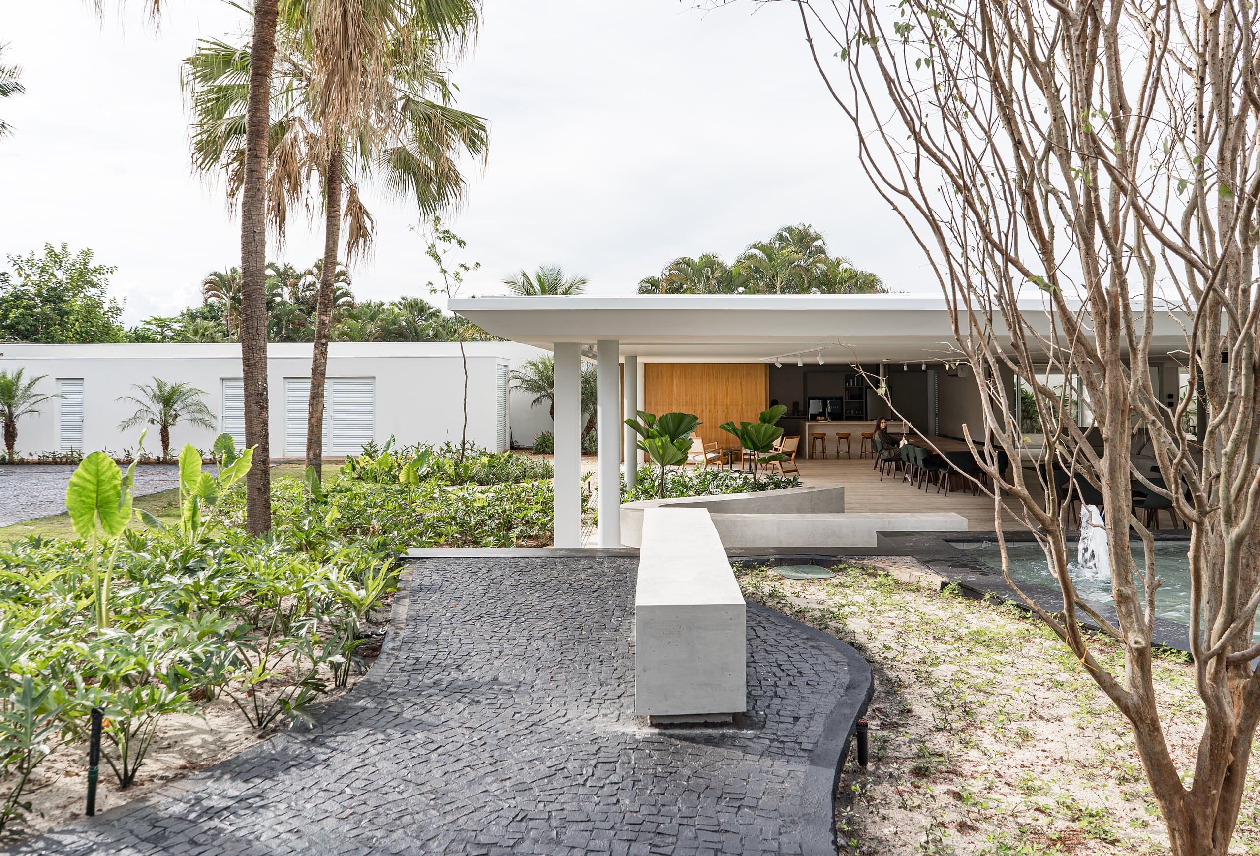 A dark cobbled path curves through tropical planting and palms towards a white flat-roofed pavilion with a timber wall inside.