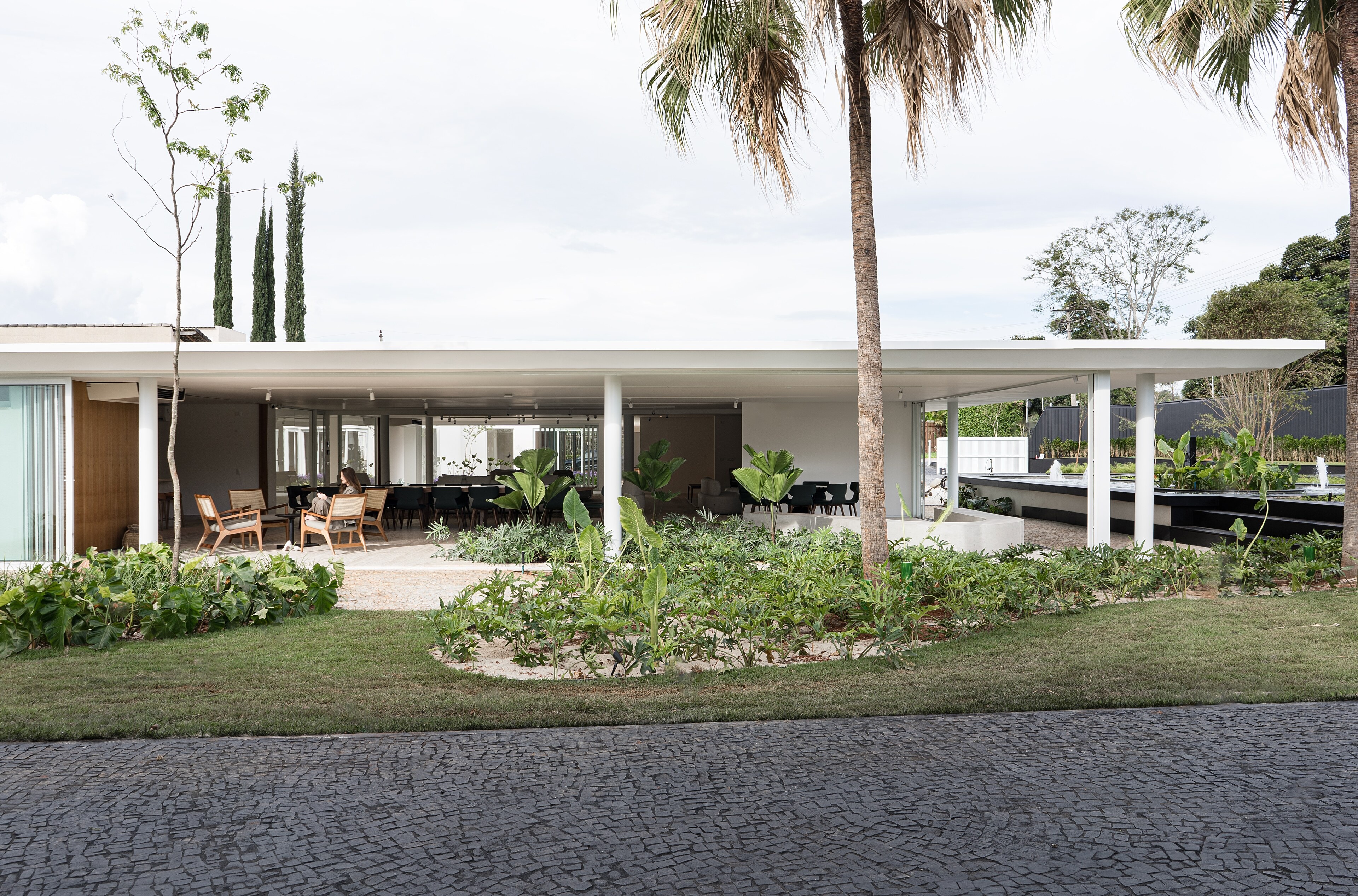 A long white pavilion under a thin flat roof, timber chairs on its open terrace, palms and a planted lawn in front.