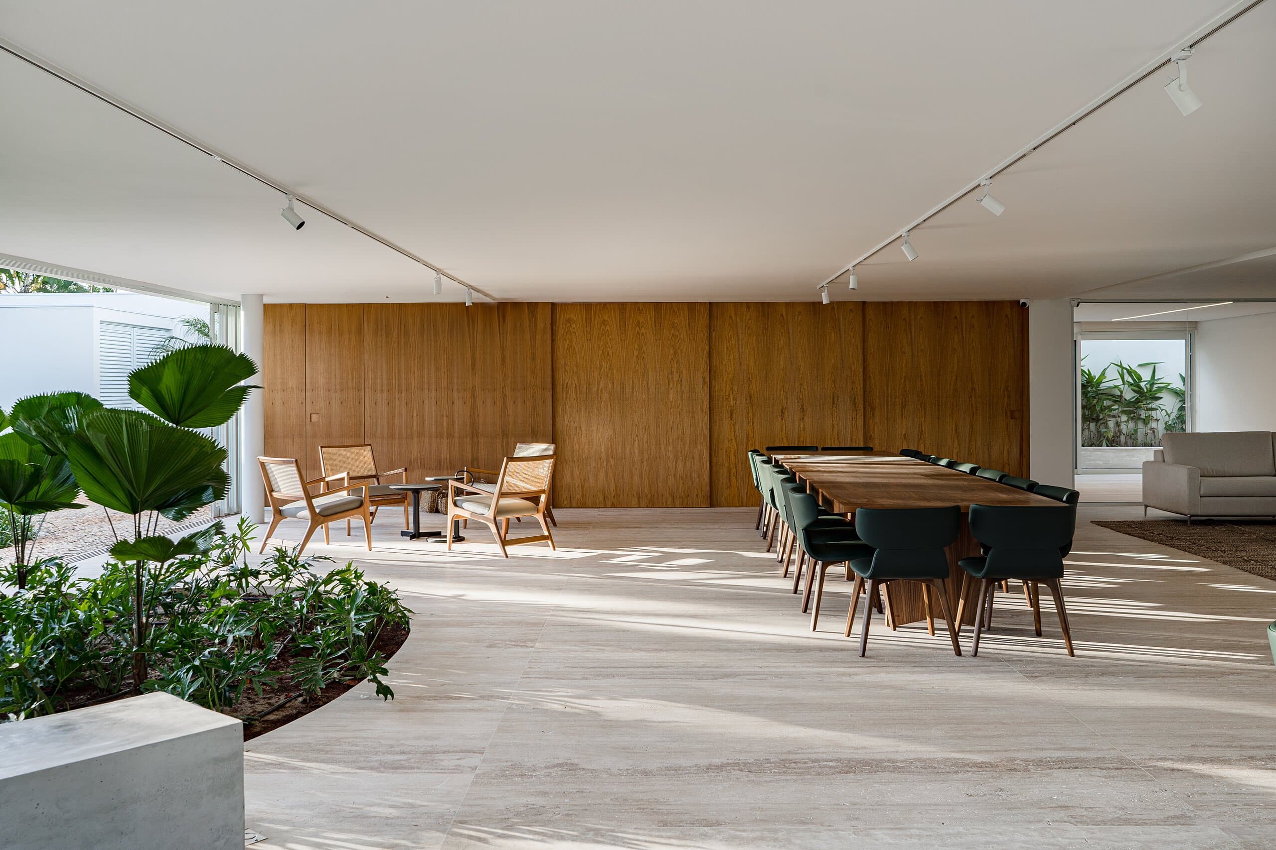 An open hall in pale stone: a long dining table with black chairs, timber lounge chairs and a full-width timber wall.