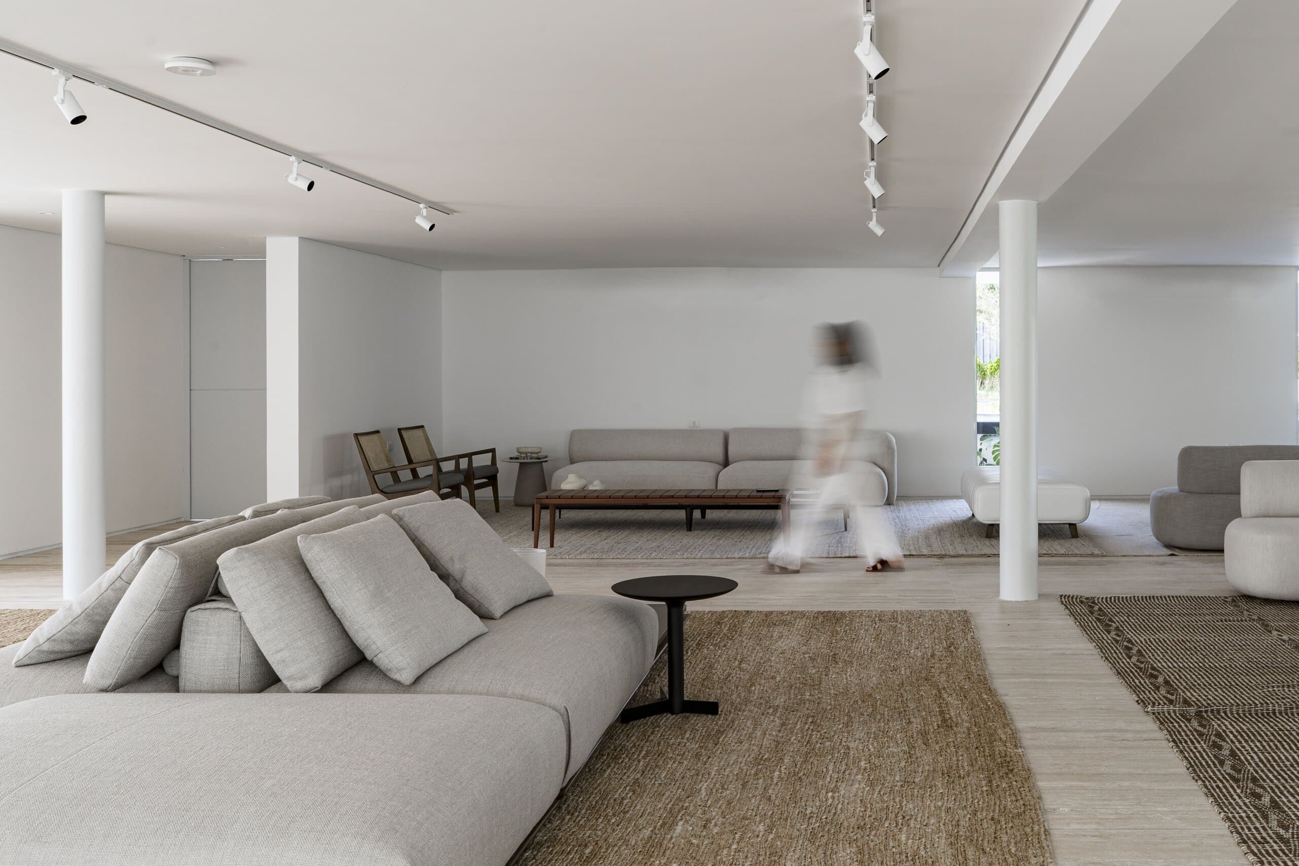 A white living room with columns and track lights: a pale sofa, a jute rug and a figure blurred in motion.
