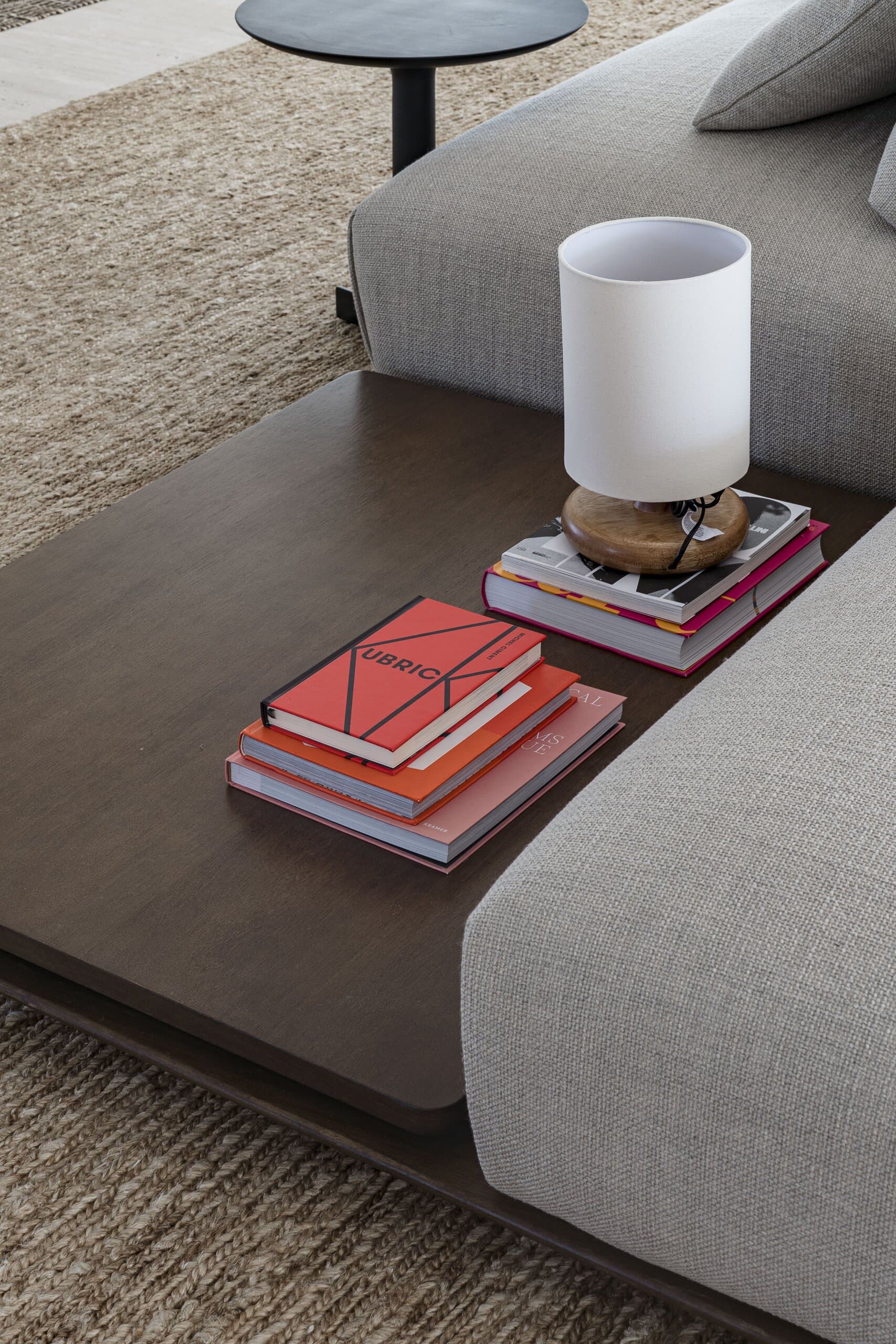 A dark timber side table between grey sofa cushions, with red books and a white lamp.