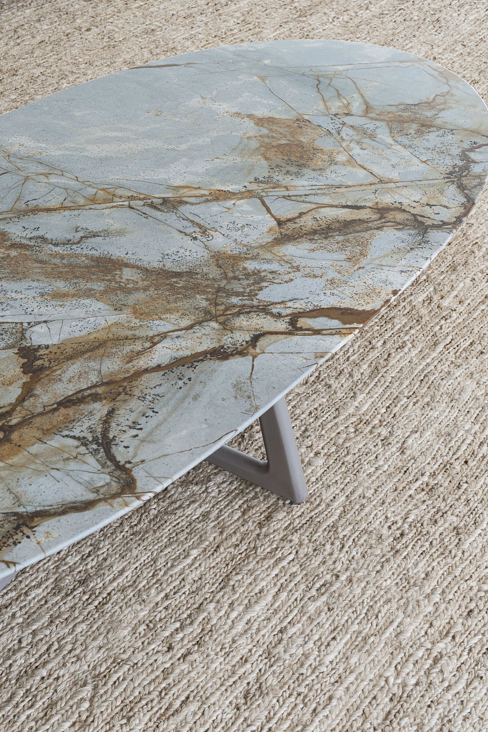 An oval stone table veined in grey and ochre on a textured jute rug.