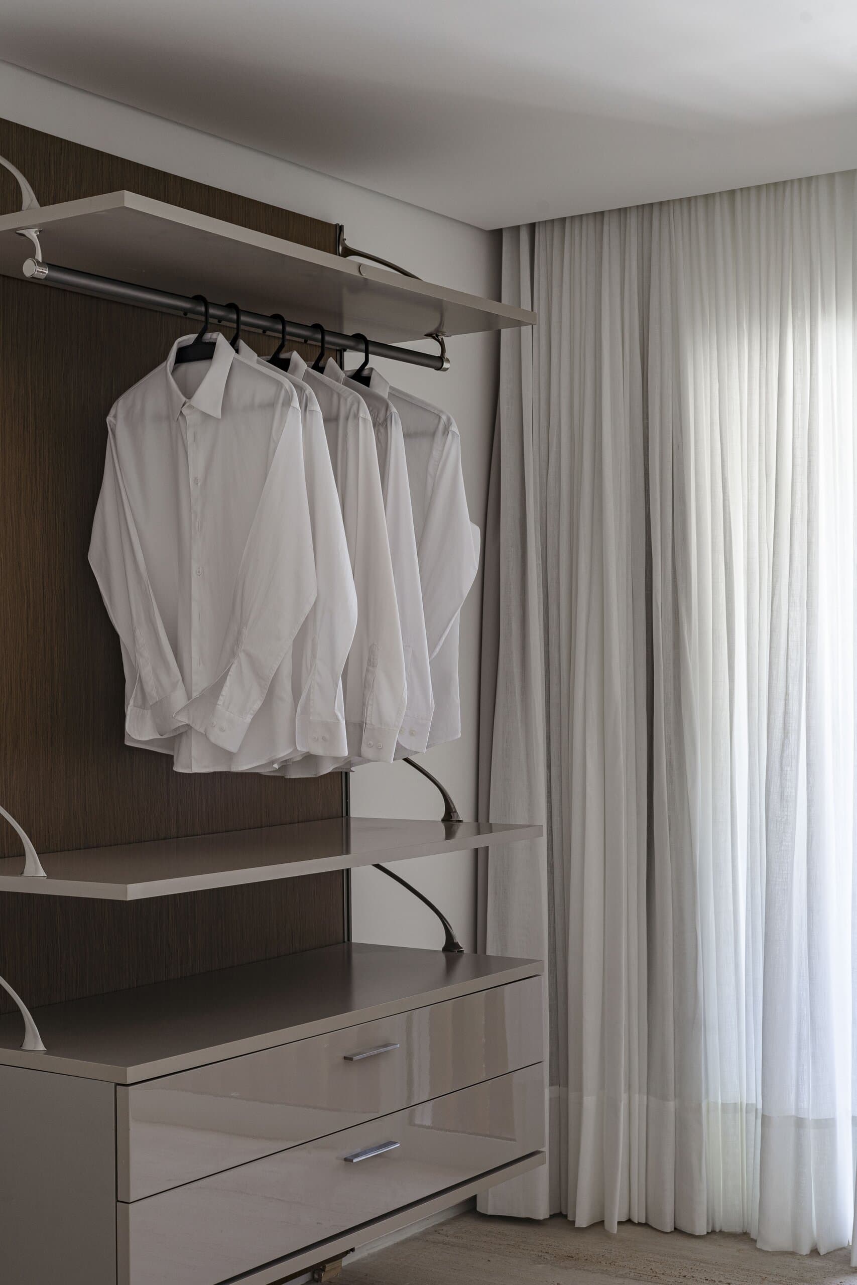White shirts on a rail in an open wardrobe beside sheer curtains.