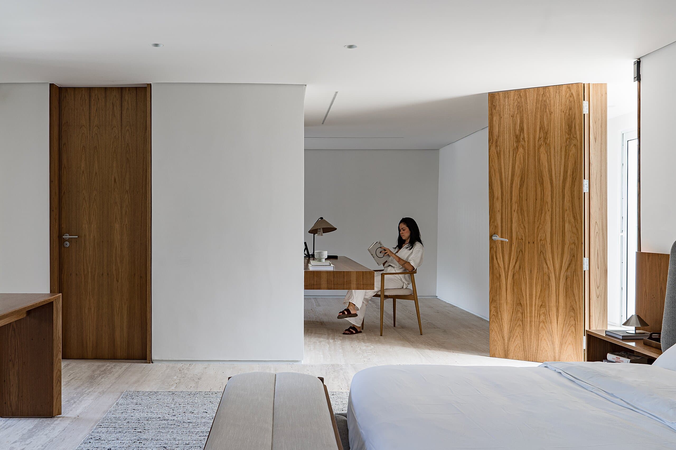 A bedroom with timber doors opening onto a study, where a woman reads at a timber desk.