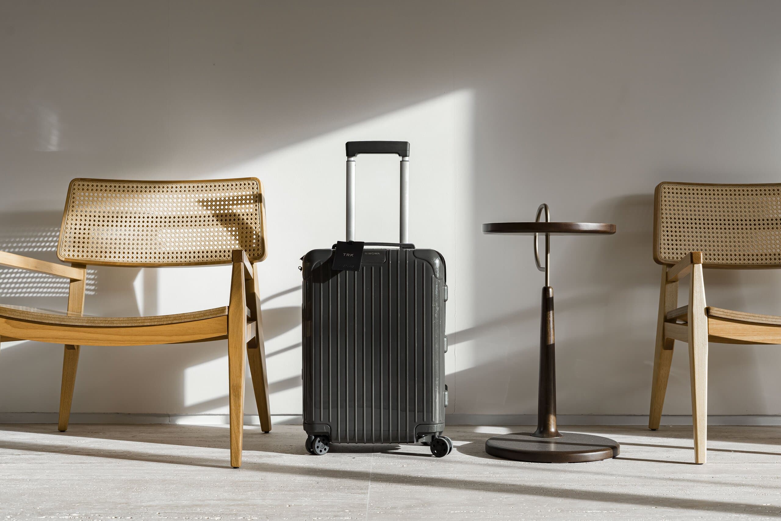 Two cane chairs, a black suitcase and a slender side table against a white wall in raking light.