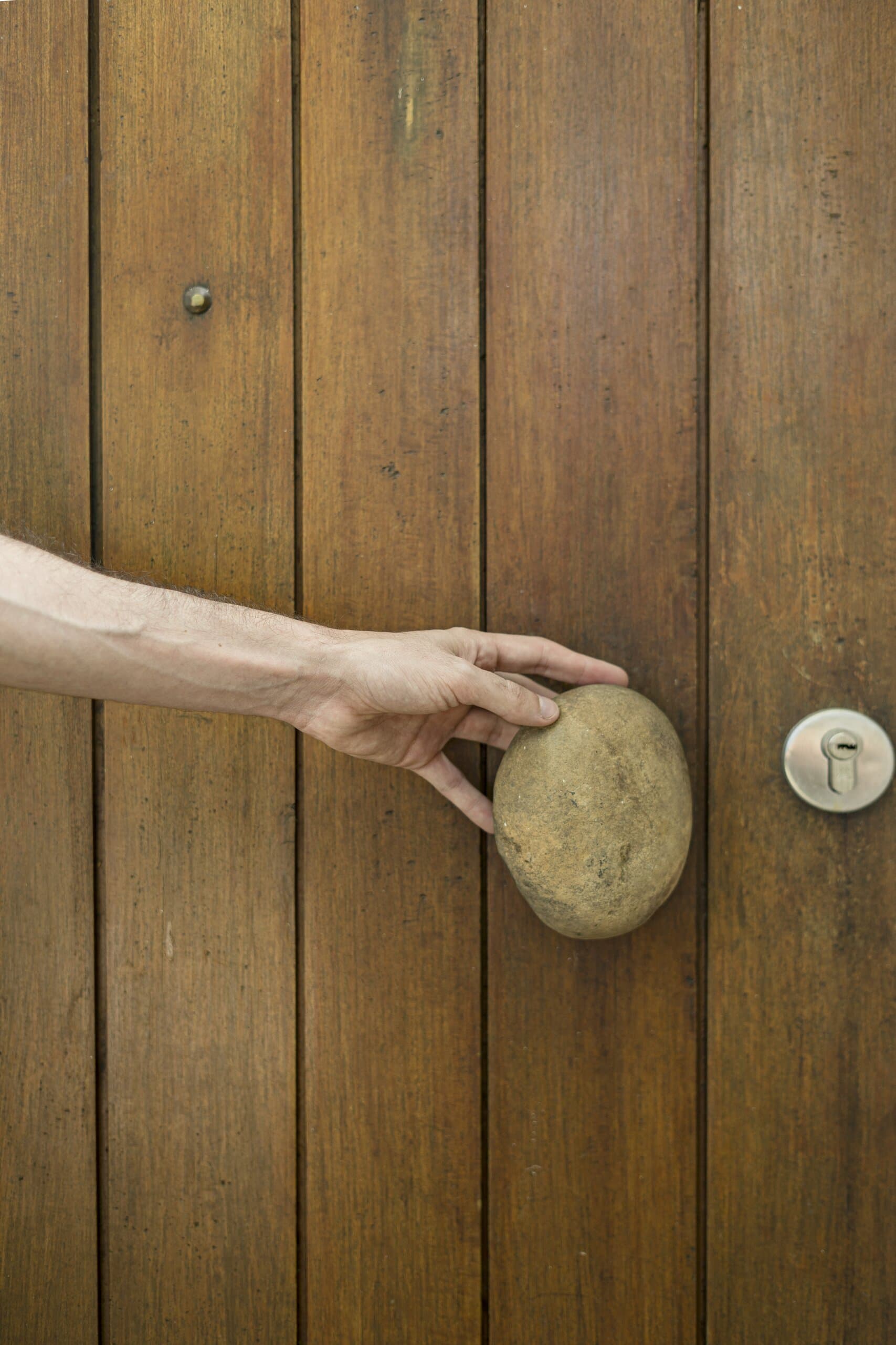 A hand grips a smooth river stone used as the handle of a timber plank door.