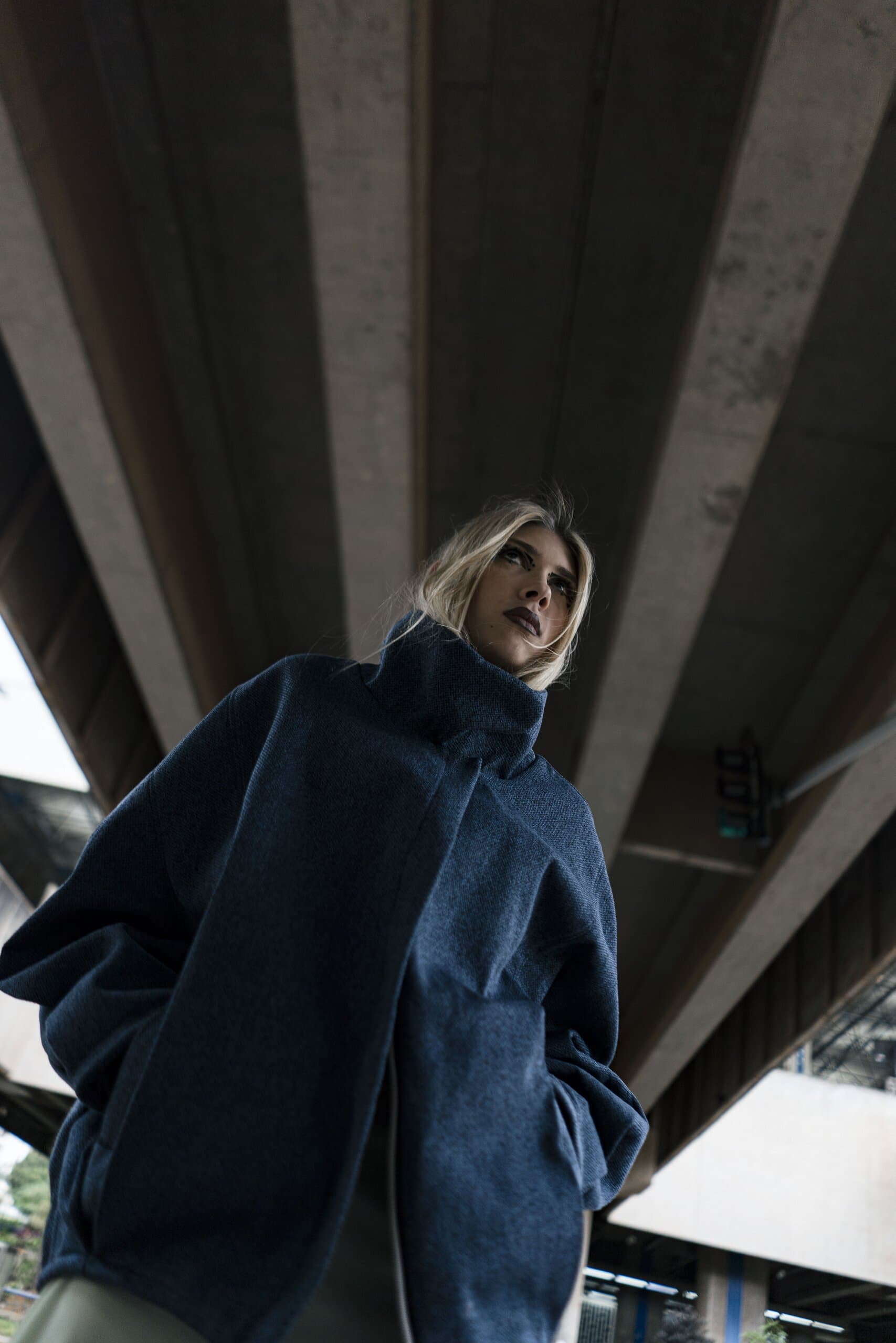 A blonde woman in an oversized navy coat looks up beneath the ribbed concrete of an overpass.