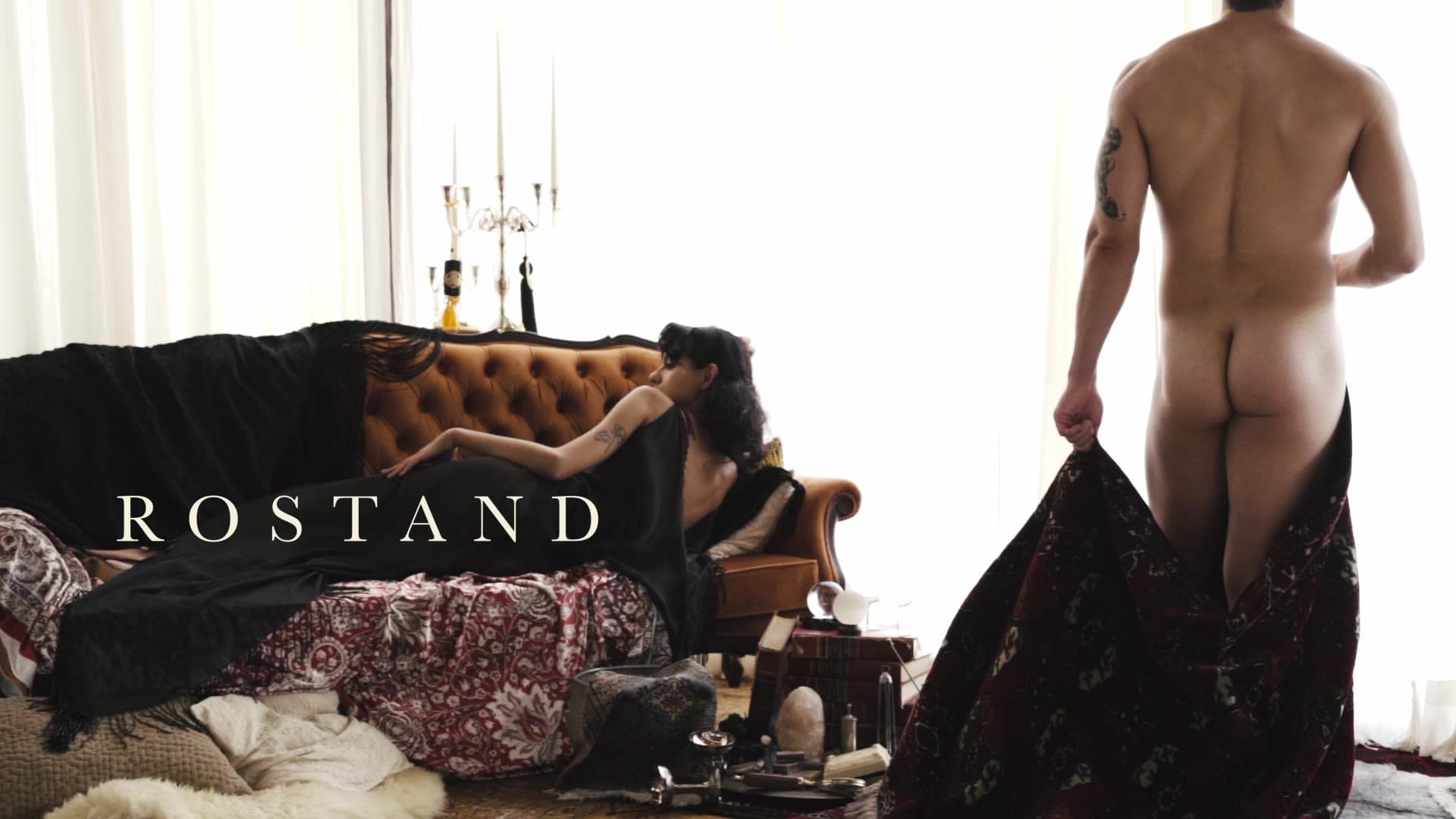 In a bright room with a candelabra and a leather sofa, a man seen from behind holds up a dark patterned cloth, the word ROSTAND across the frame.