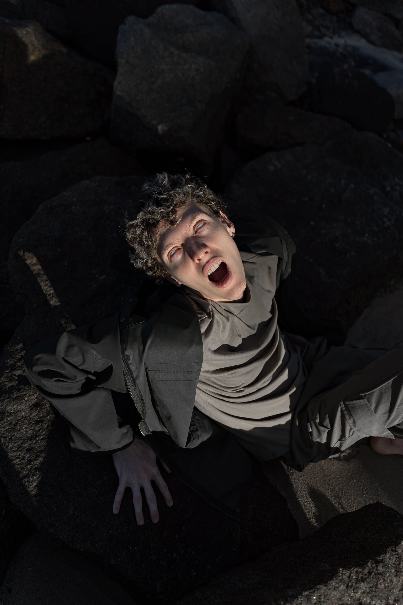 From above: a curly-haired man lies among dark rocks, mouth open in a shout, a hard shaft of light across his face.
