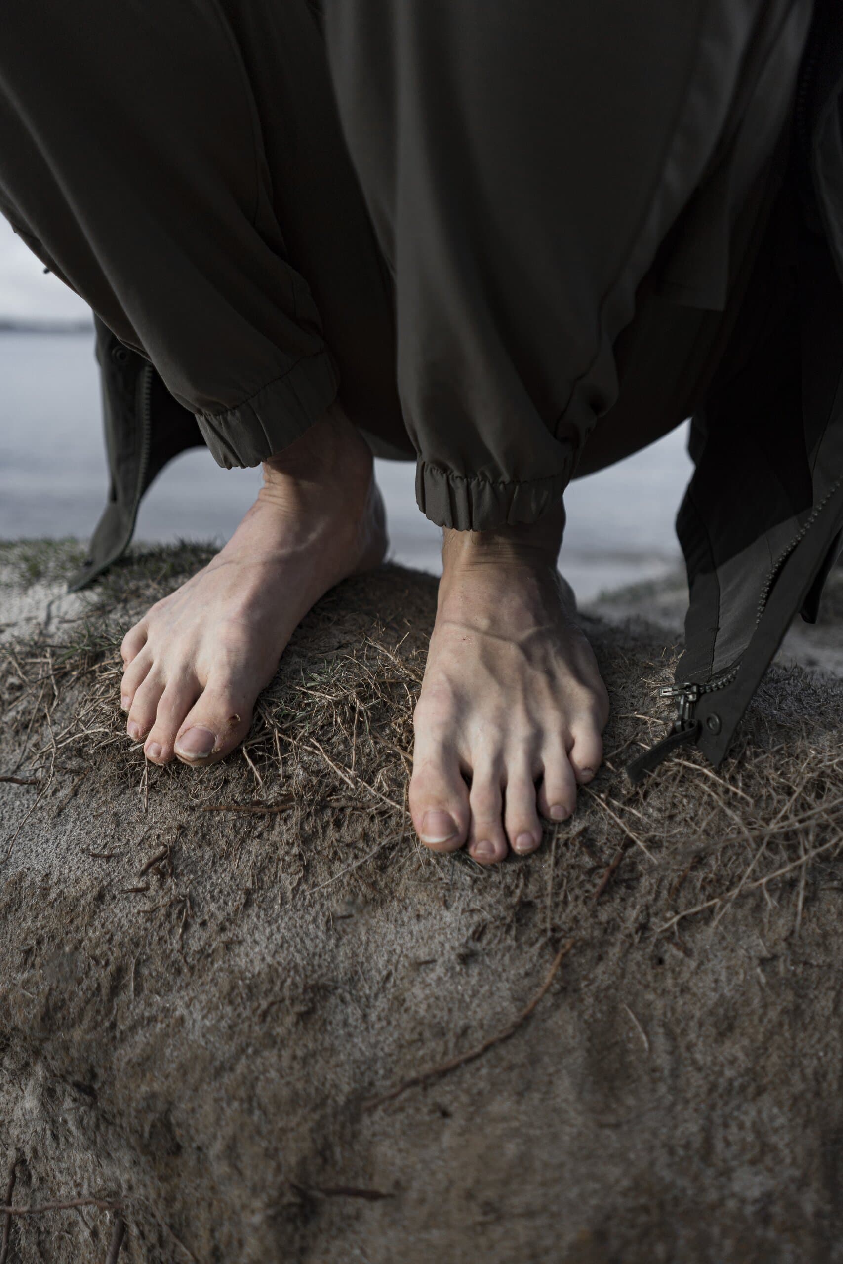 Bare feet on a crumbling earth ledge, the hem of loose dark trousers and a coat gathered above.