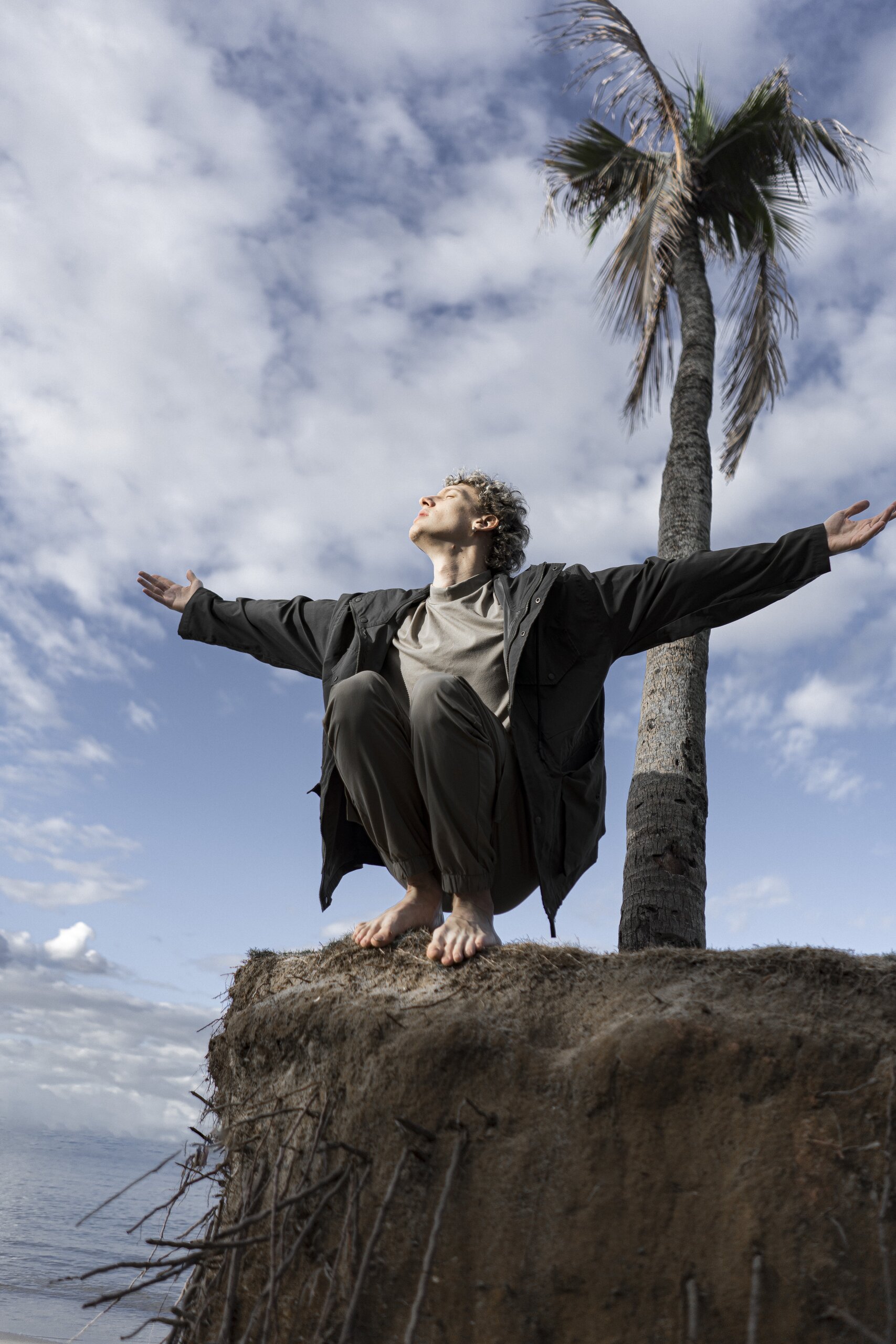 A young man in a dark coat crouches barefoot on the edge of an eroded earth bank, arms spread wide beneath a cloudy blue sky and a palm tree.