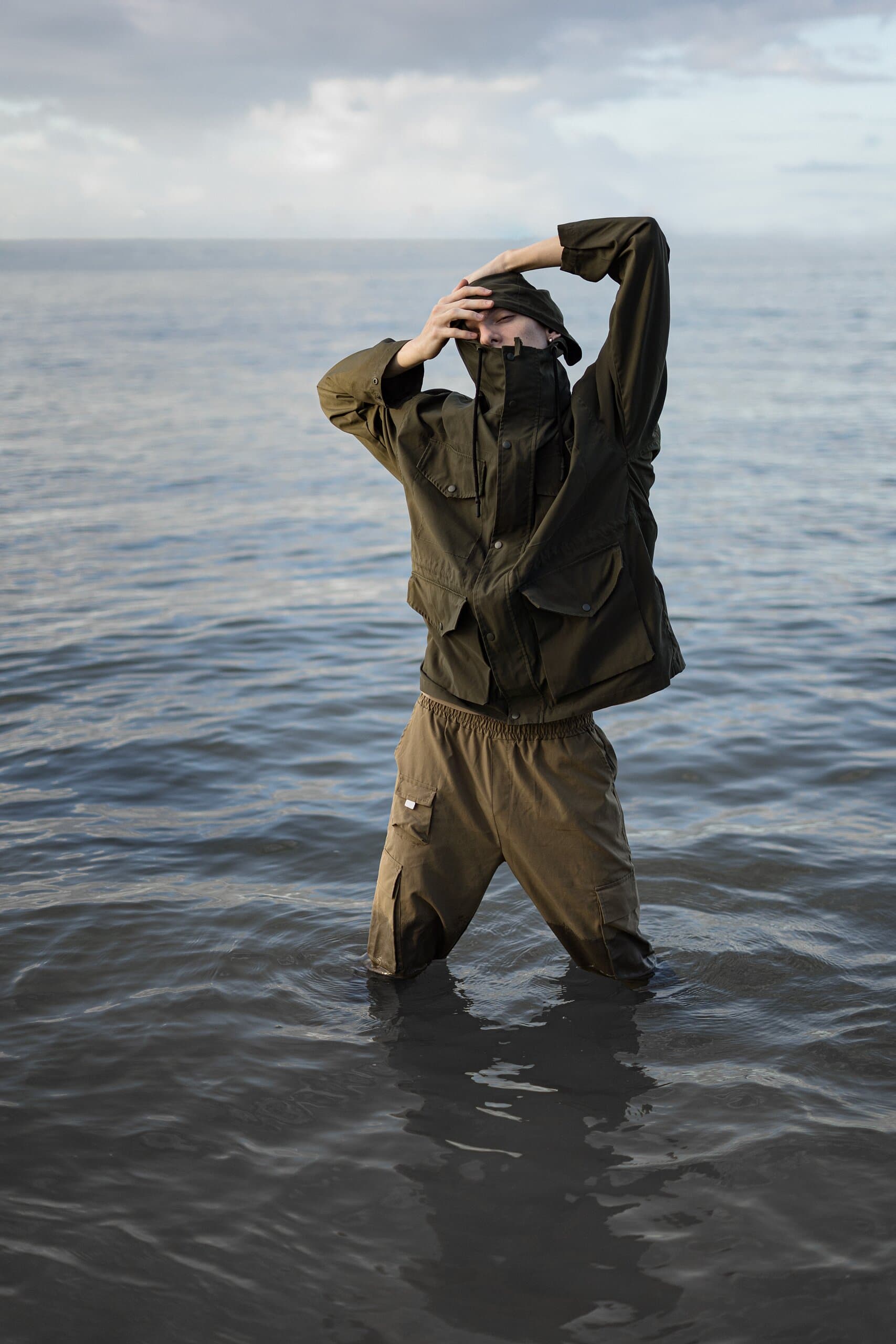 Waist-deep in calm grey sea, a man in an olive jacket covers his face with both arms.