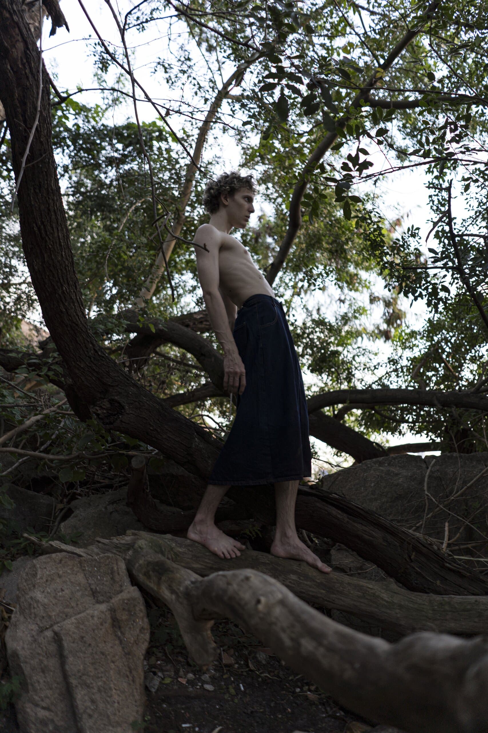 A shirtless man in a long denim skirt stands barefoot on twisted roots beneath a leaning tree by the water.