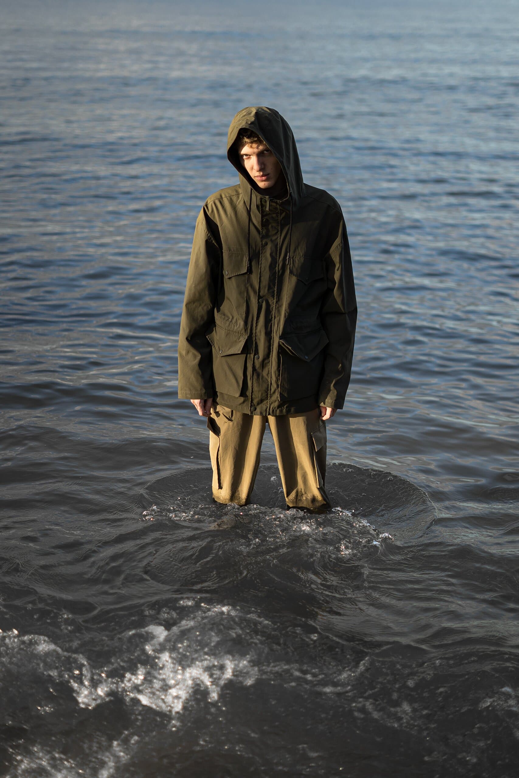 A hooded figure in an olive parka stands knee-deep in the sea, a small wave breaking at his legs.