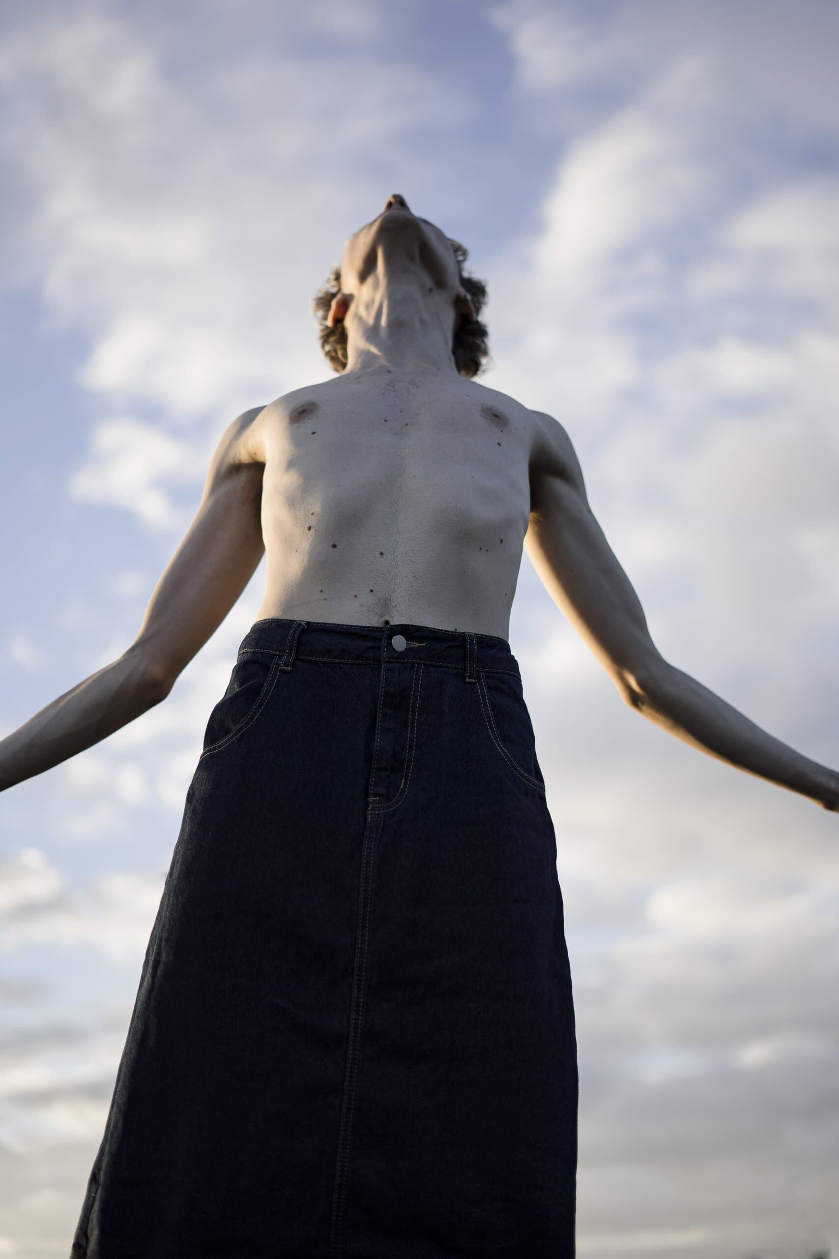 From below: a shirtless man in a denim skirt throws his head back, arms open against a sky of white clouds.