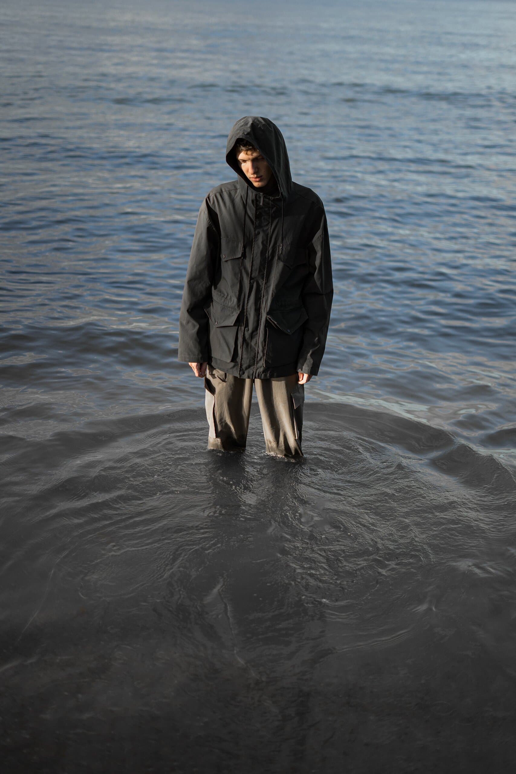 The hooded figure in a dark parka stands still in the sea, ripples spreading around his legs.