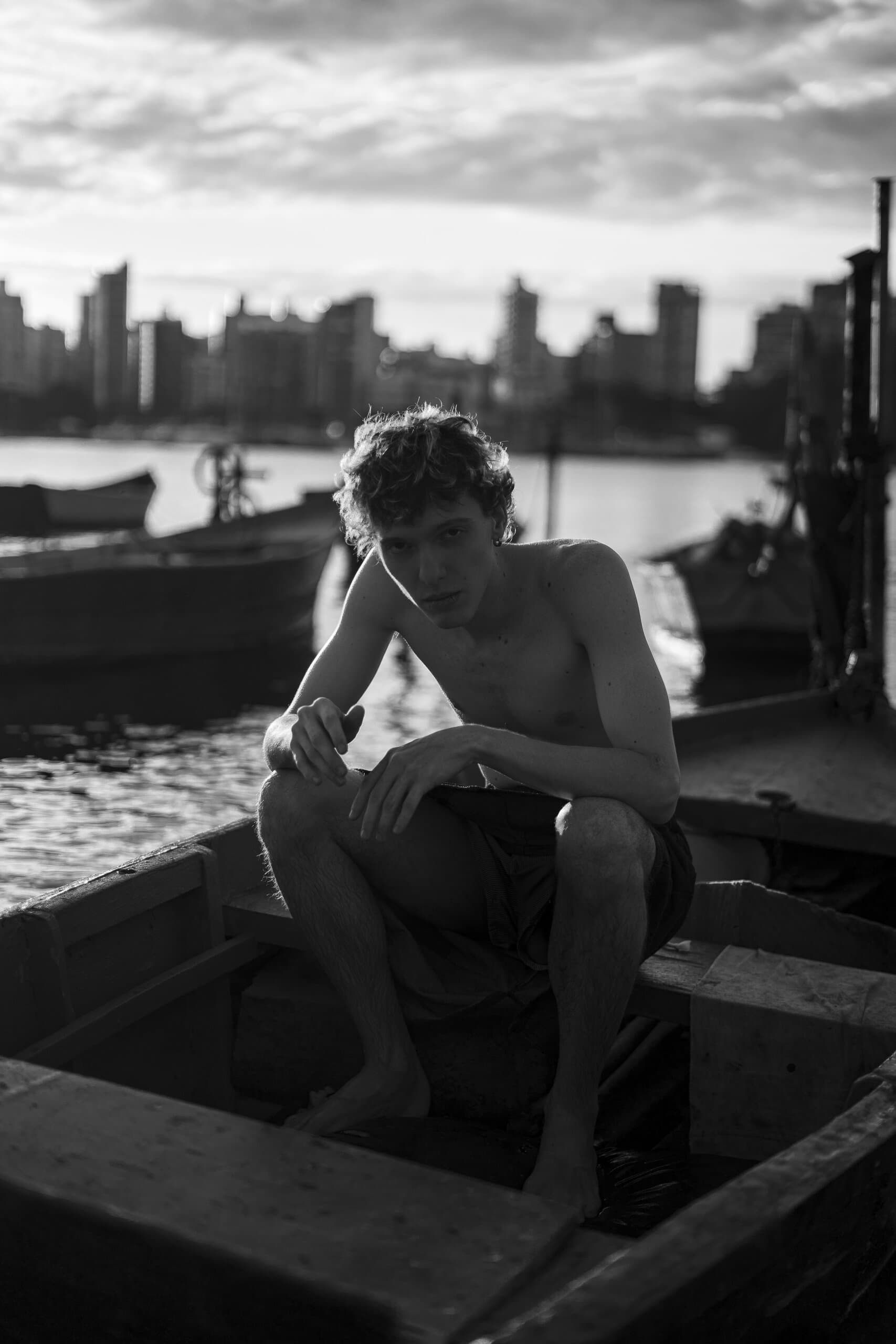 In black and white: a shirtless man crouches in a wooden fishing boat, a line of tower blocks across the bay behind him.