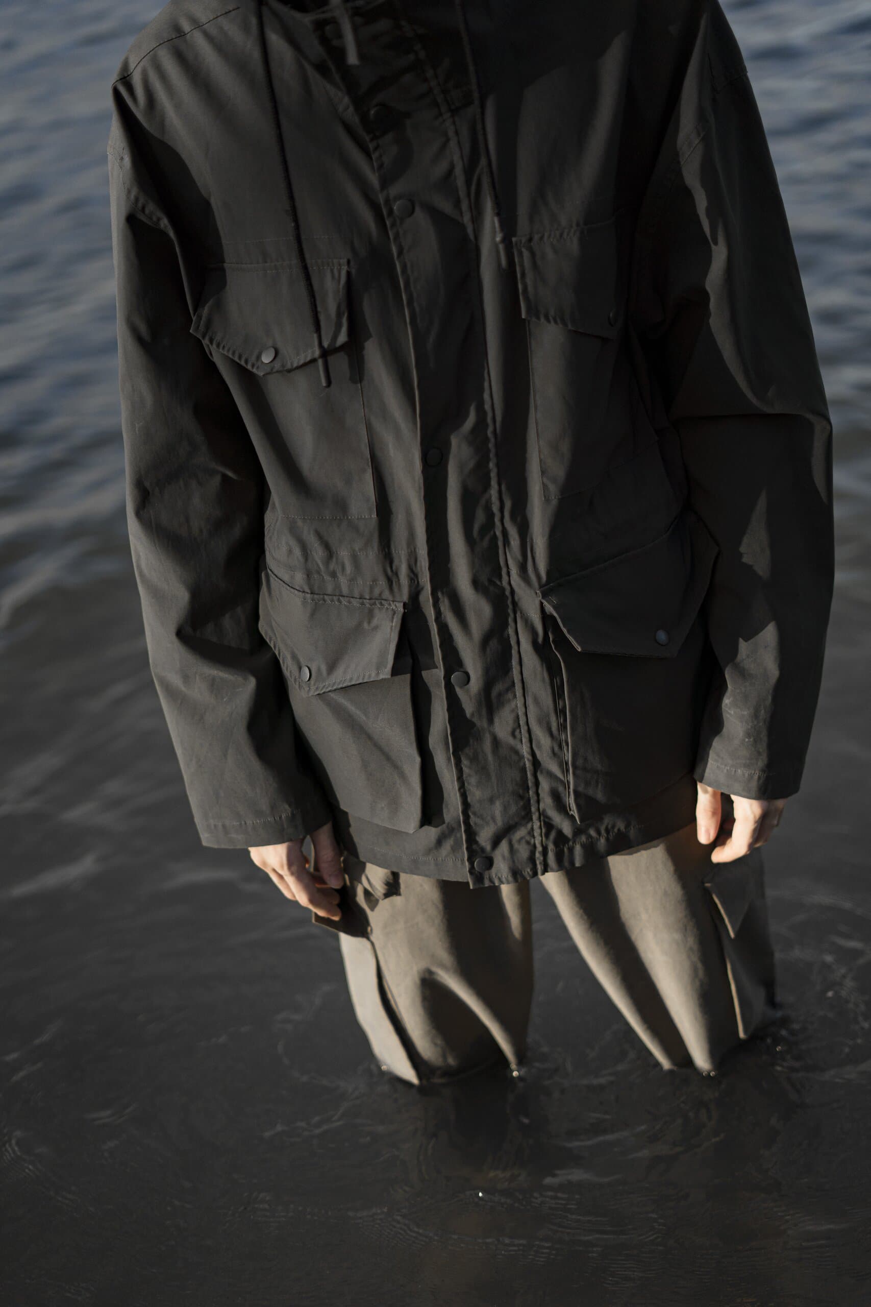 Close on a dark parka and grey trousers, hands hanging at the sides, legs disappearing into the water.