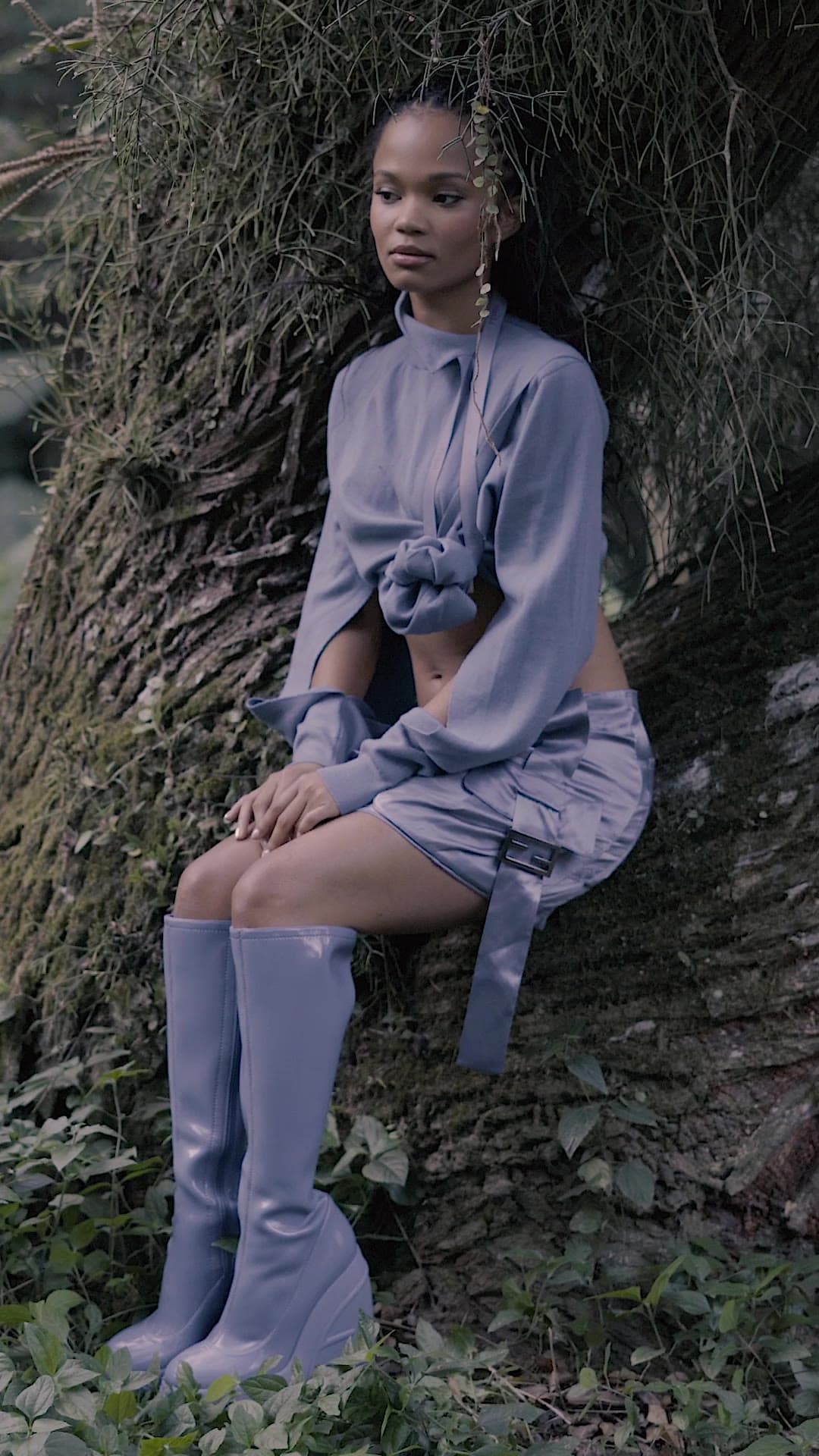A woman in a lilac cropped jacket, mini skirt and knee-high boots sits against a moss-covered tree trunk.