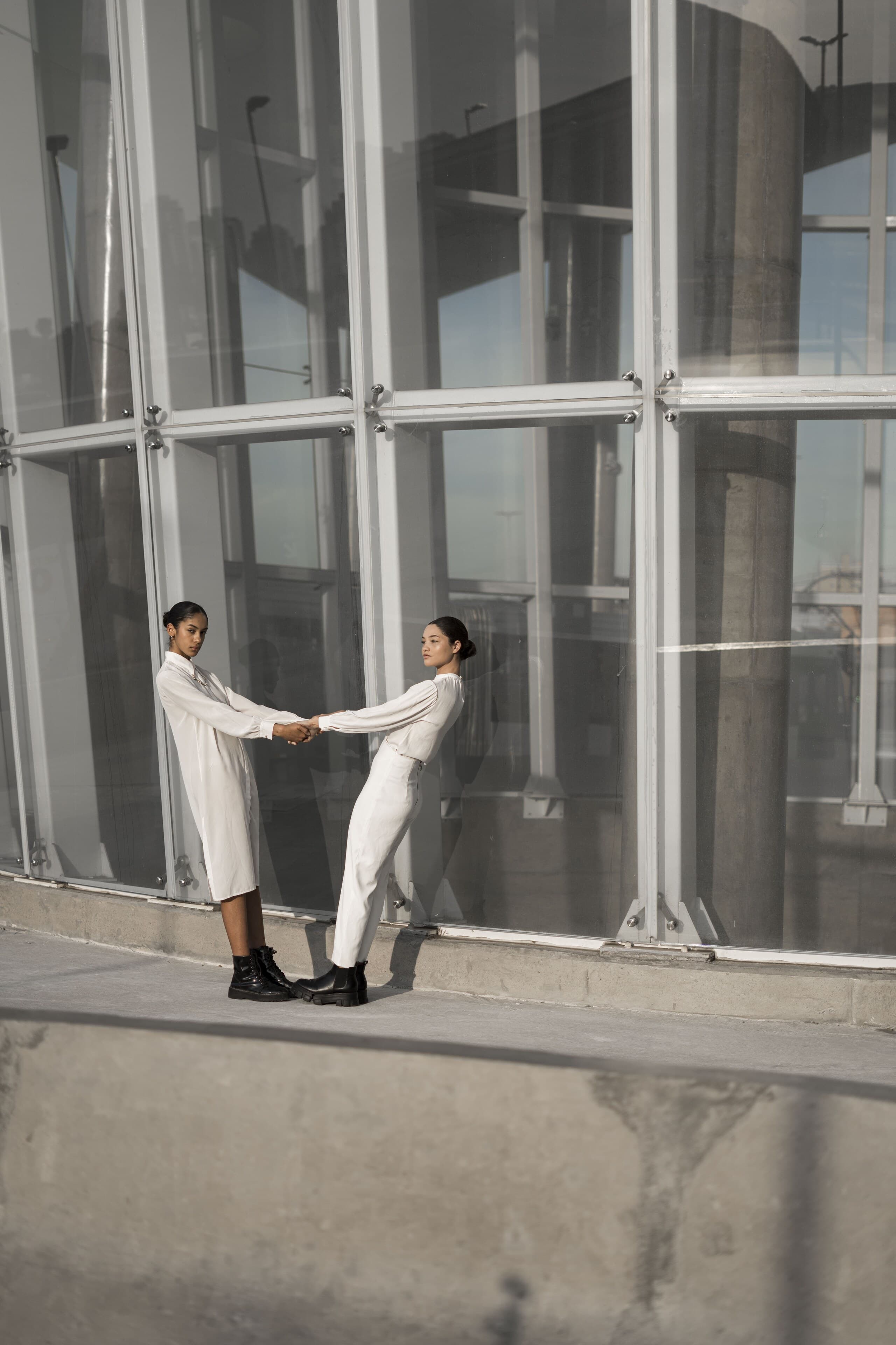 Two women in white hold hands at arm's length in front of a tall steel-framed glass facade.