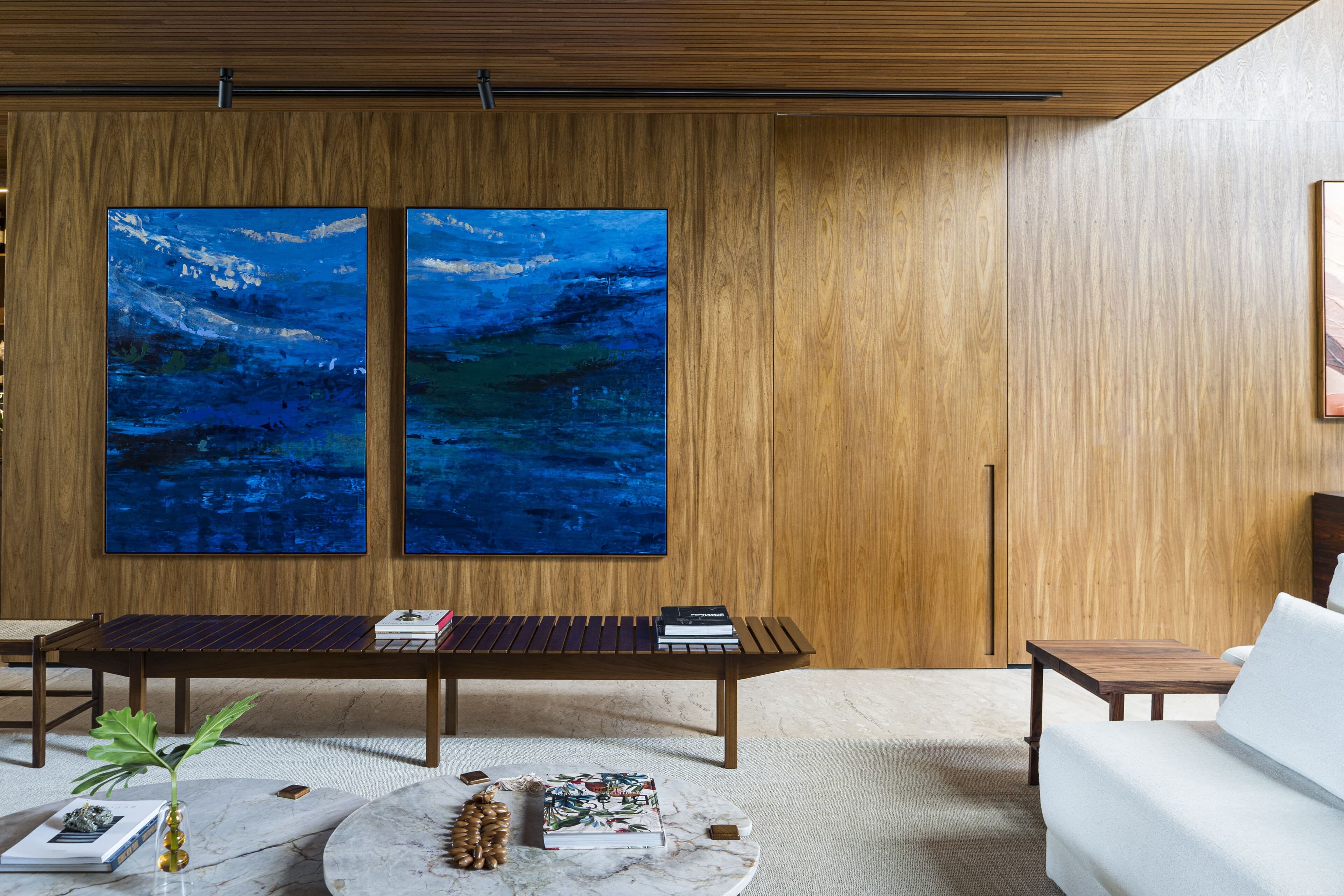 Two deep-blue paintings above a slatted timber bench, a round marble table in front.