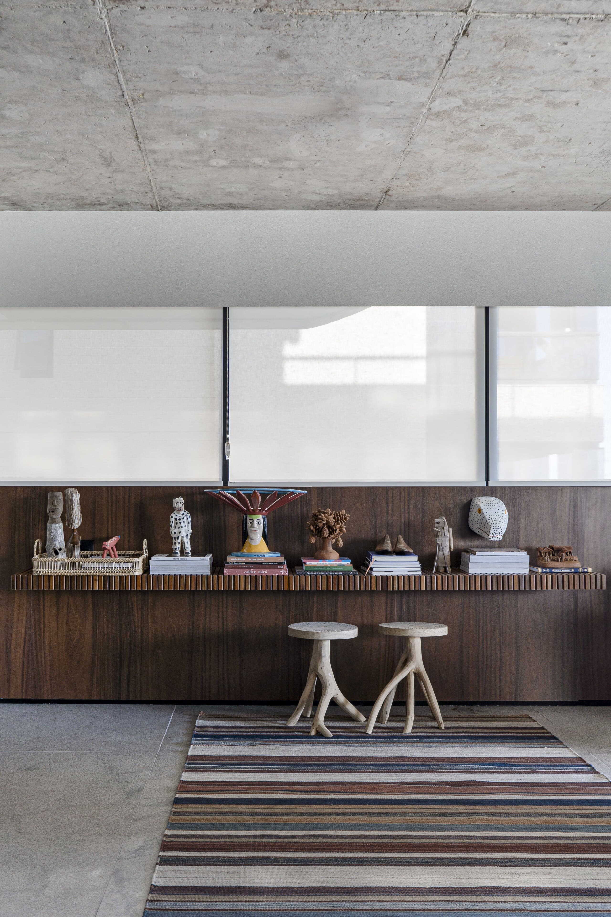 Beneath a raw concrete ceiling, a timber console of objects and two sculptural stools on a striped rug.