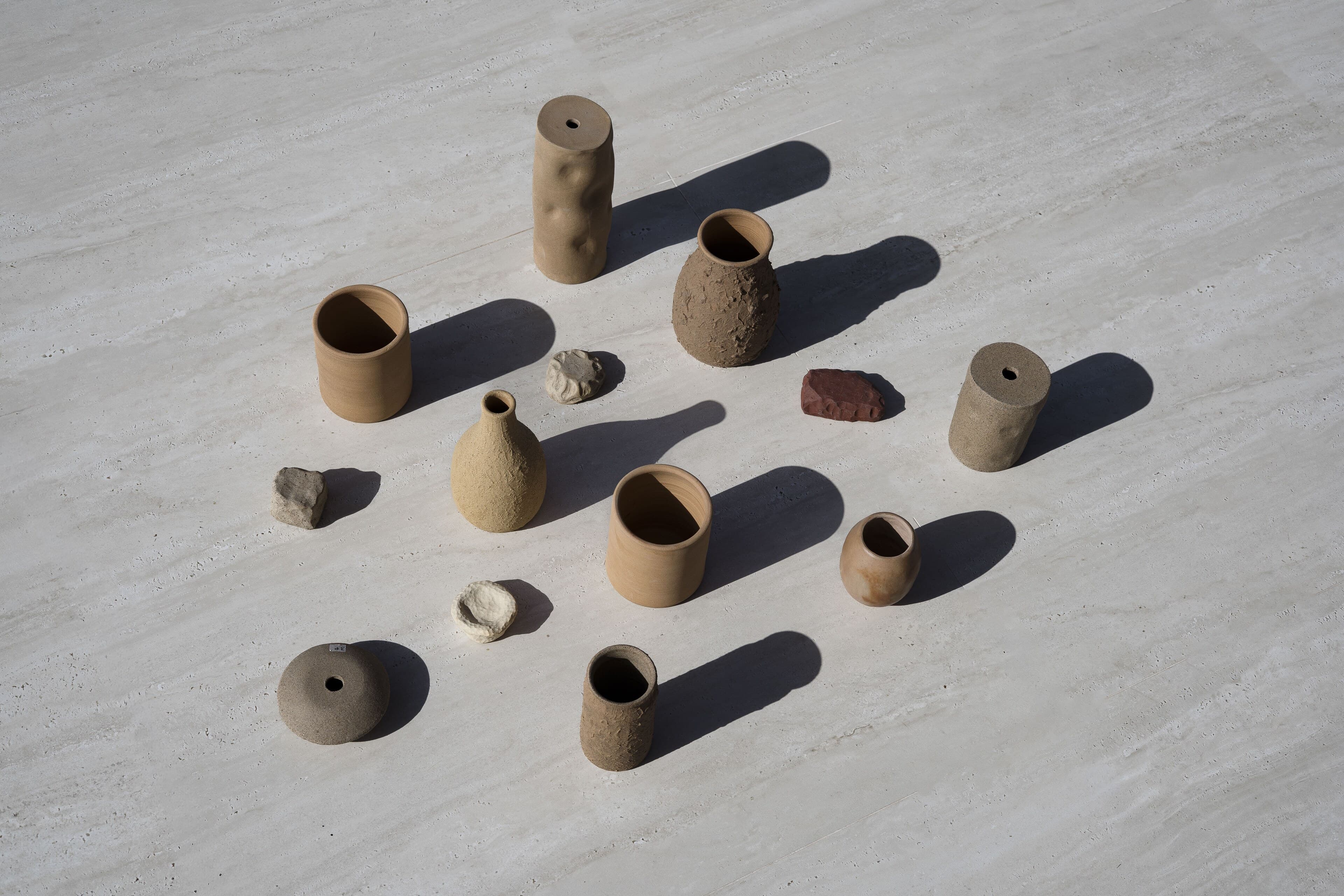 From above: hand-built ceramic vessels and small stones scattered across pale travertine, each with a long shadow.