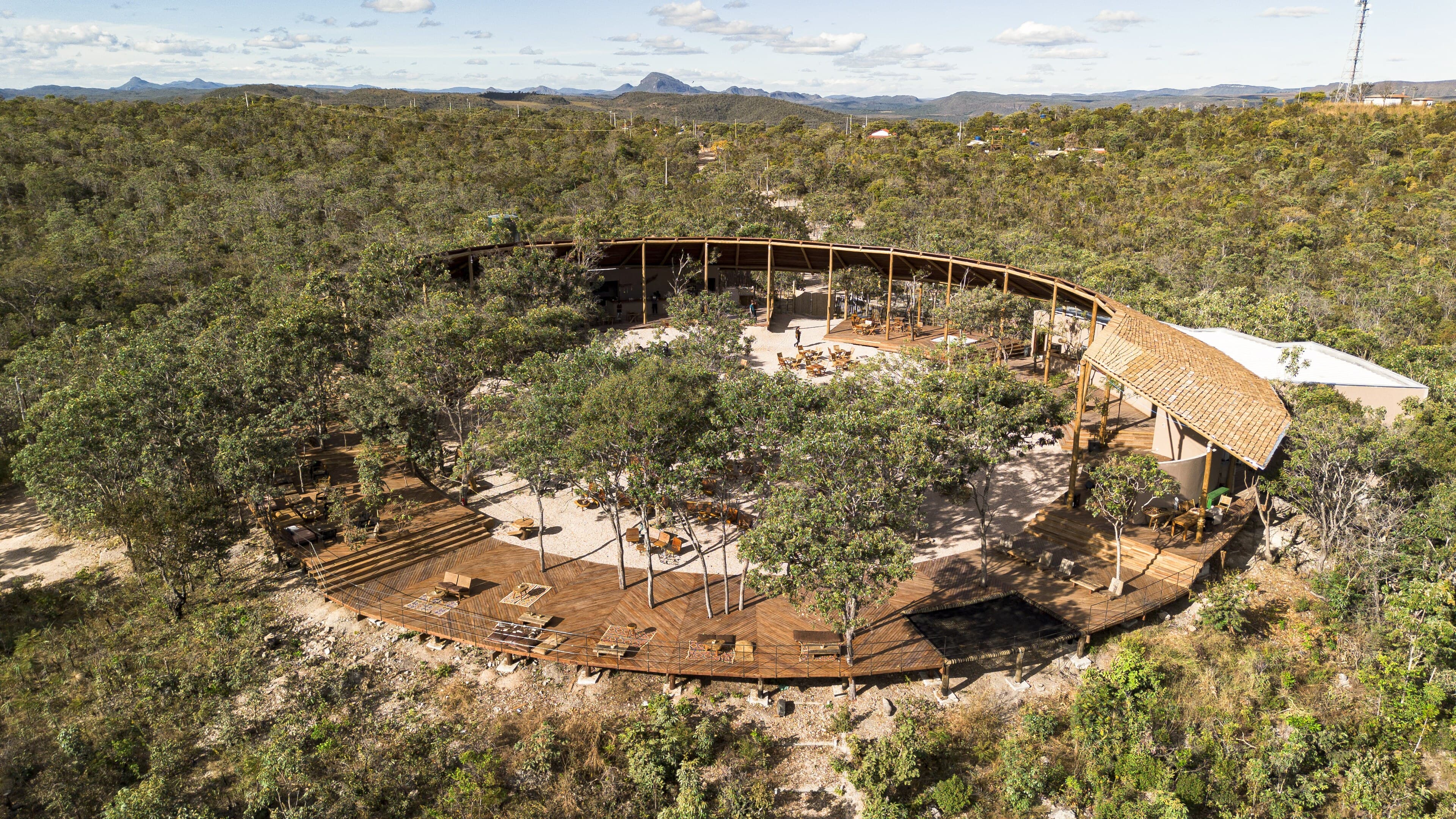 From the air: a ring of thatched roof around an open courtyard of trees, set on a rocky hilltop in the scrubland.