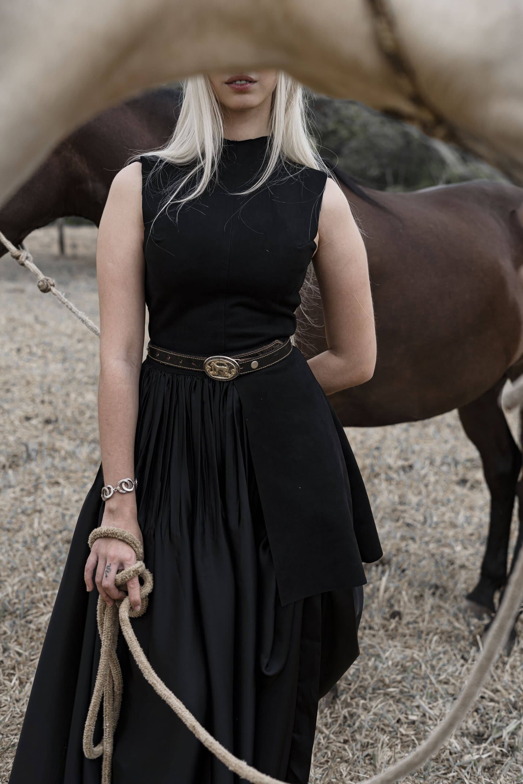 Framed under a horse's neck, a woman in a black sleeveless dress and silver-buckled belt holds a coiled rope.