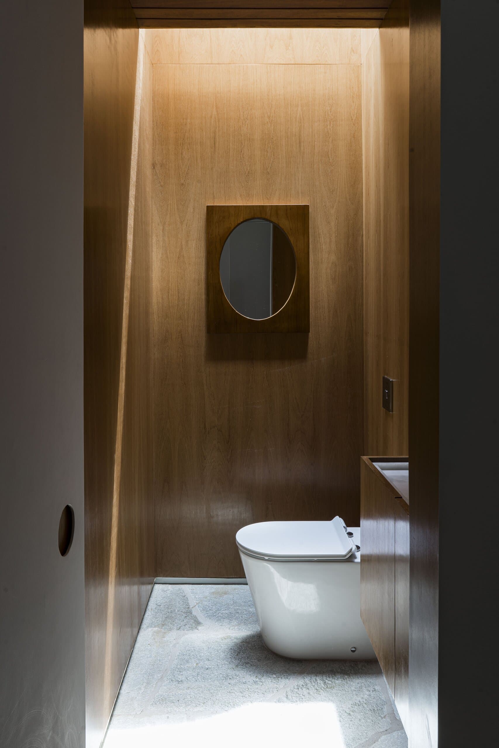 A washroom lined in timber, a round mirror in a wooden frame and a white toilet on a stone floor under a slot of light.